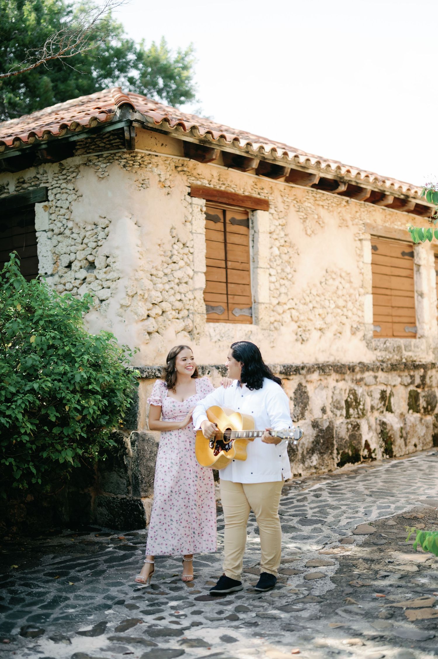 Musicians perform with a guitar against a rustic stone wall building with Spanish tile roof in a romantic garden setting.