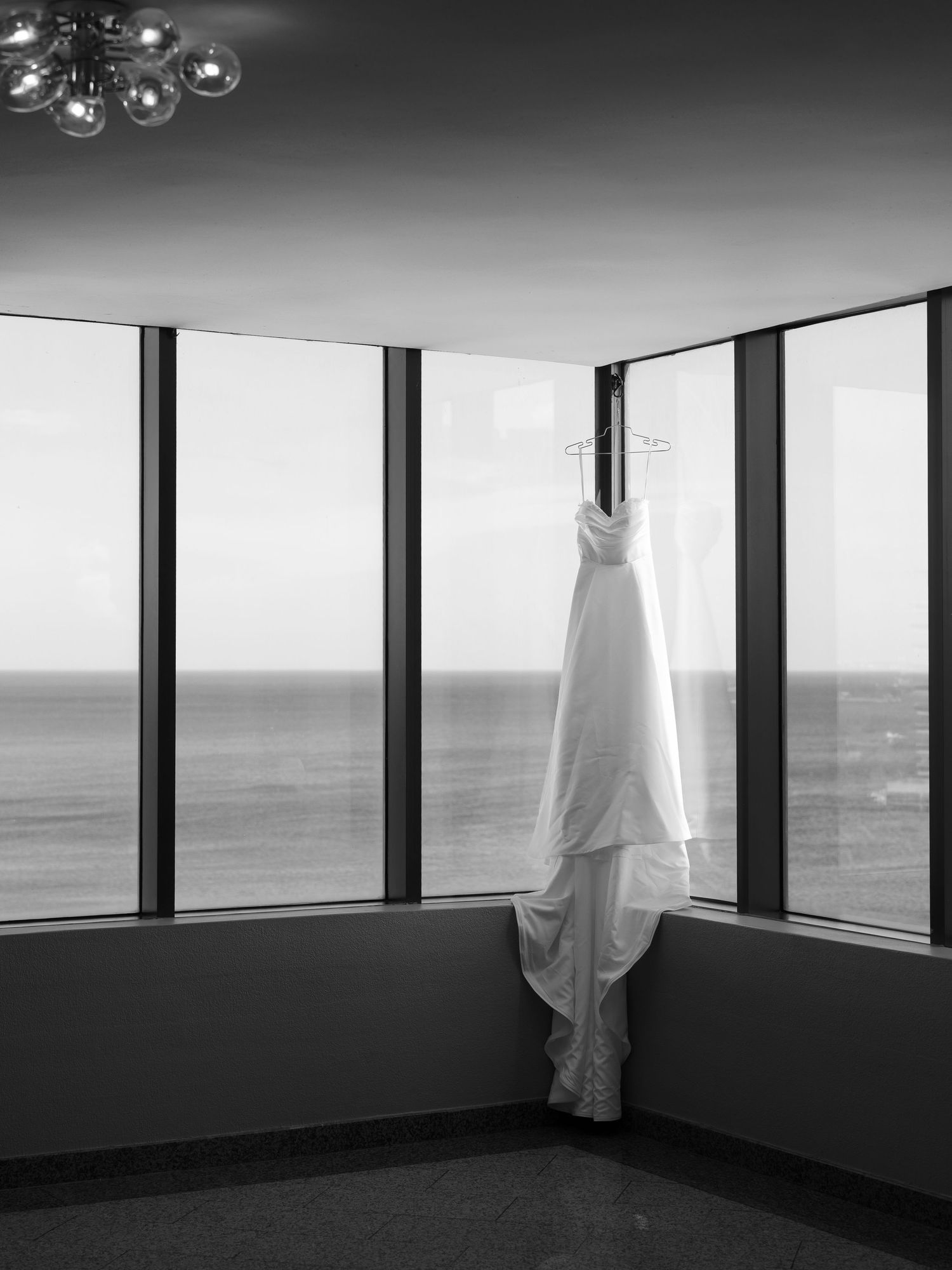 A white wedding dress hangs elegantly in a modern room with floor-to-ceiling windows overlooking the ocean.
