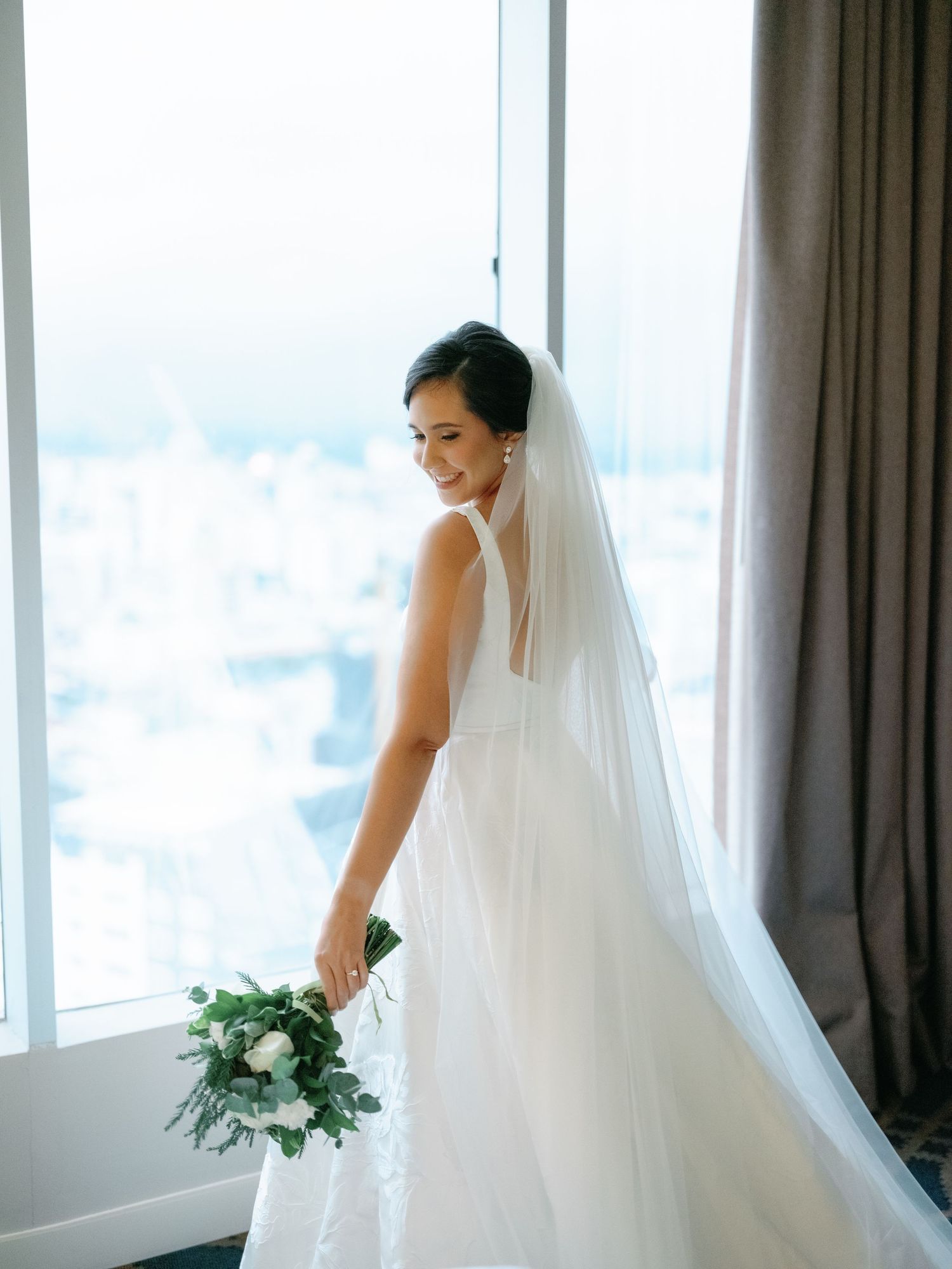 A bride in a flowing white wedding dress and veil stands elegantly near large windows with natural light.