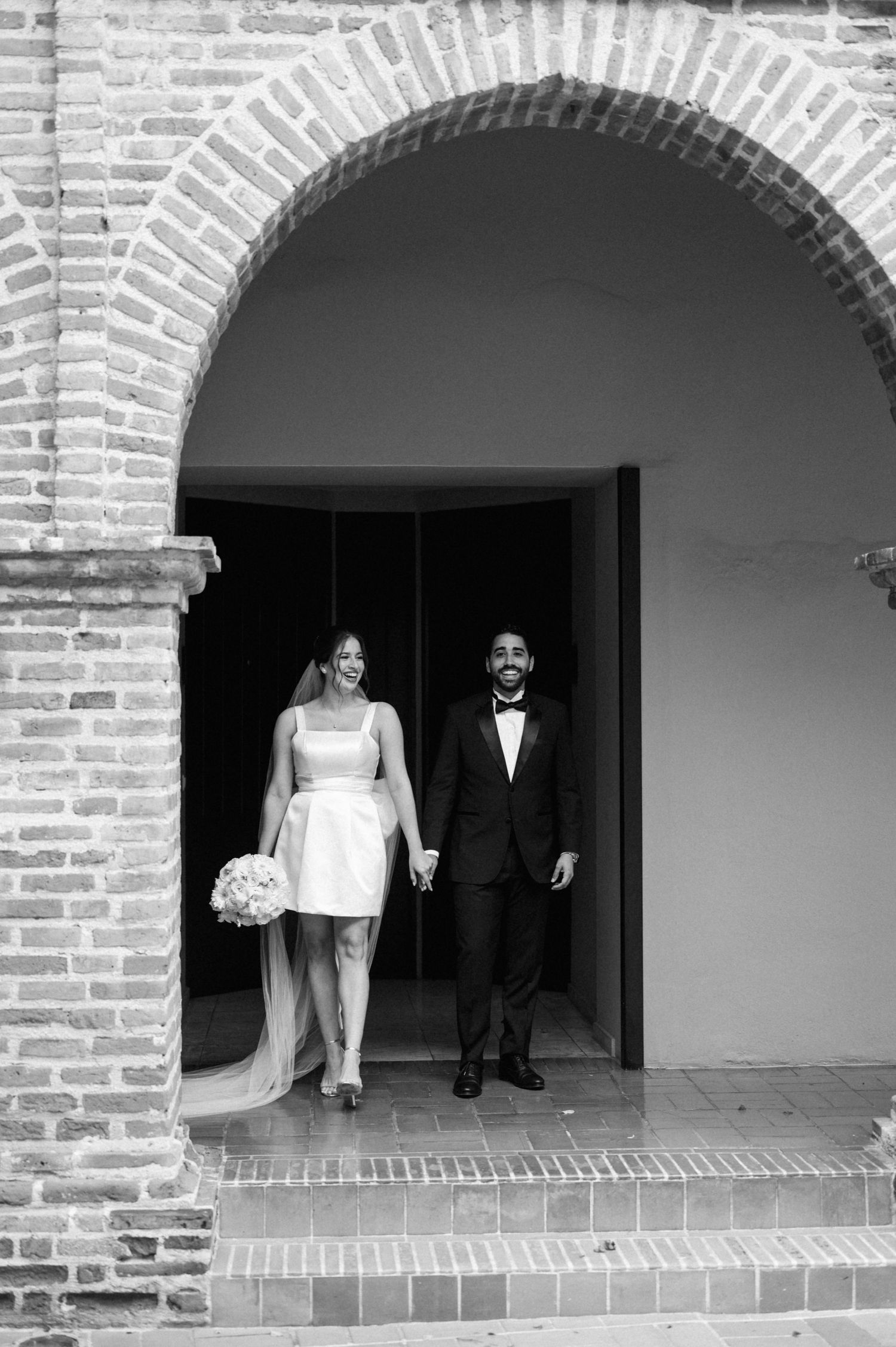 A couple in wedding attire stands under a brick archway in black and white photography.
