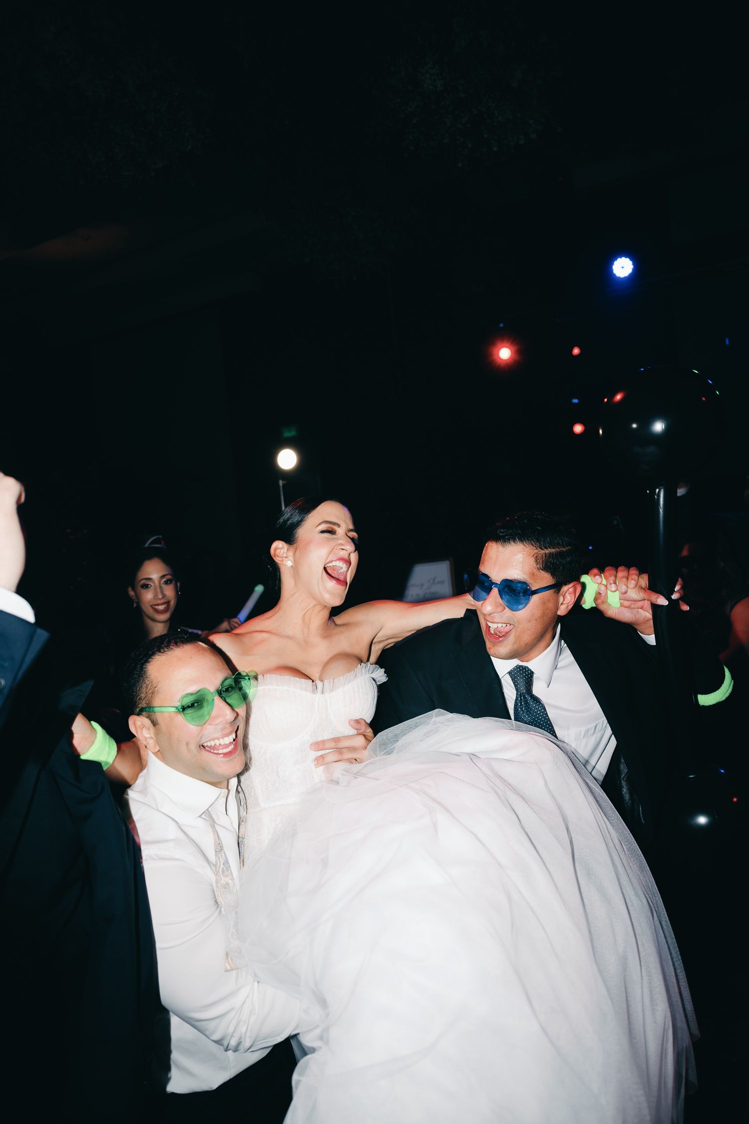 People dancing and celebrating at a wedding reception with the bride in a white gown and guests wearing sunglasses.
