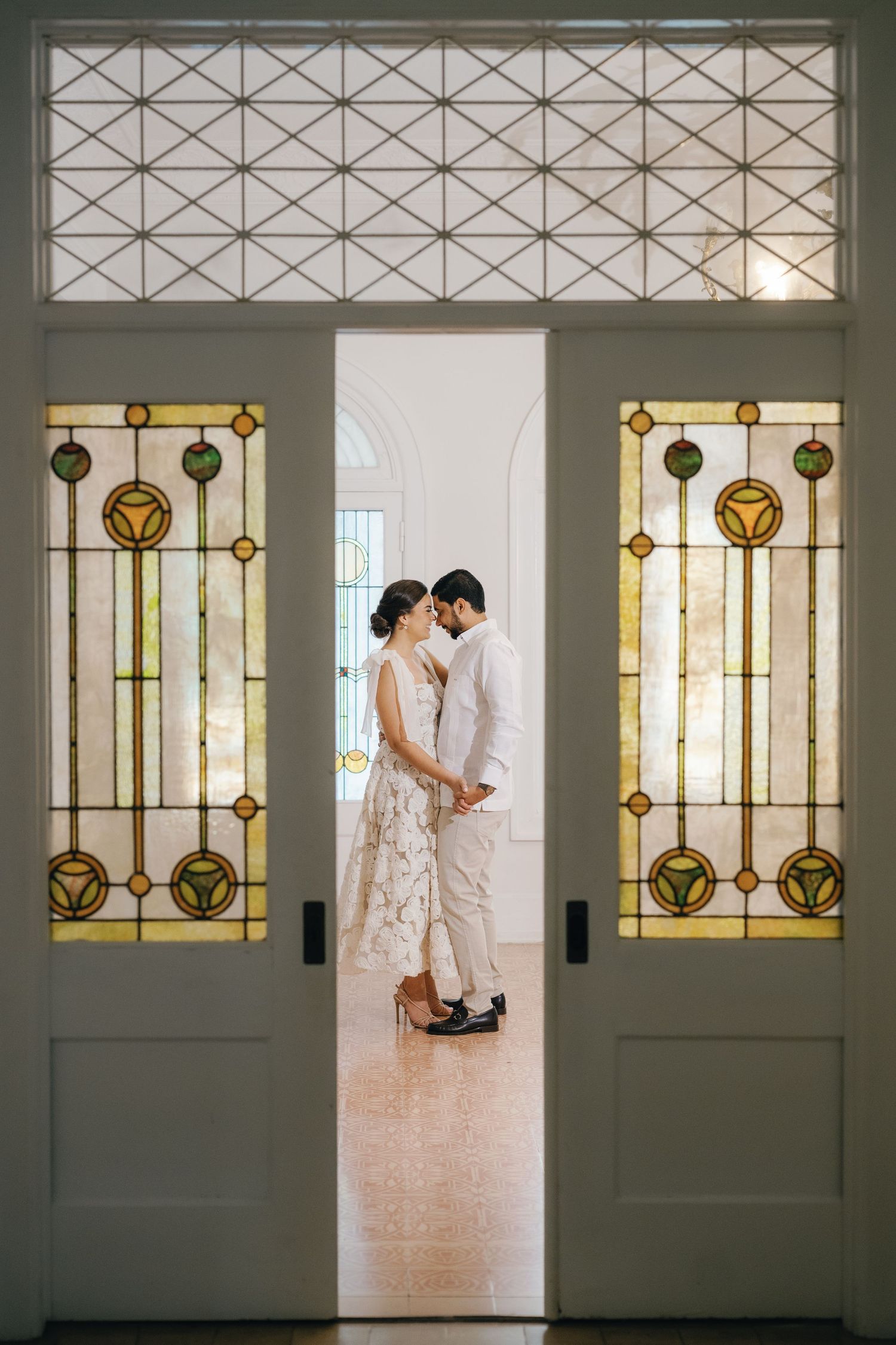 Elegant stained glass windows with geometric patterns frame a romantic moment in a bright, minimalist interior space.