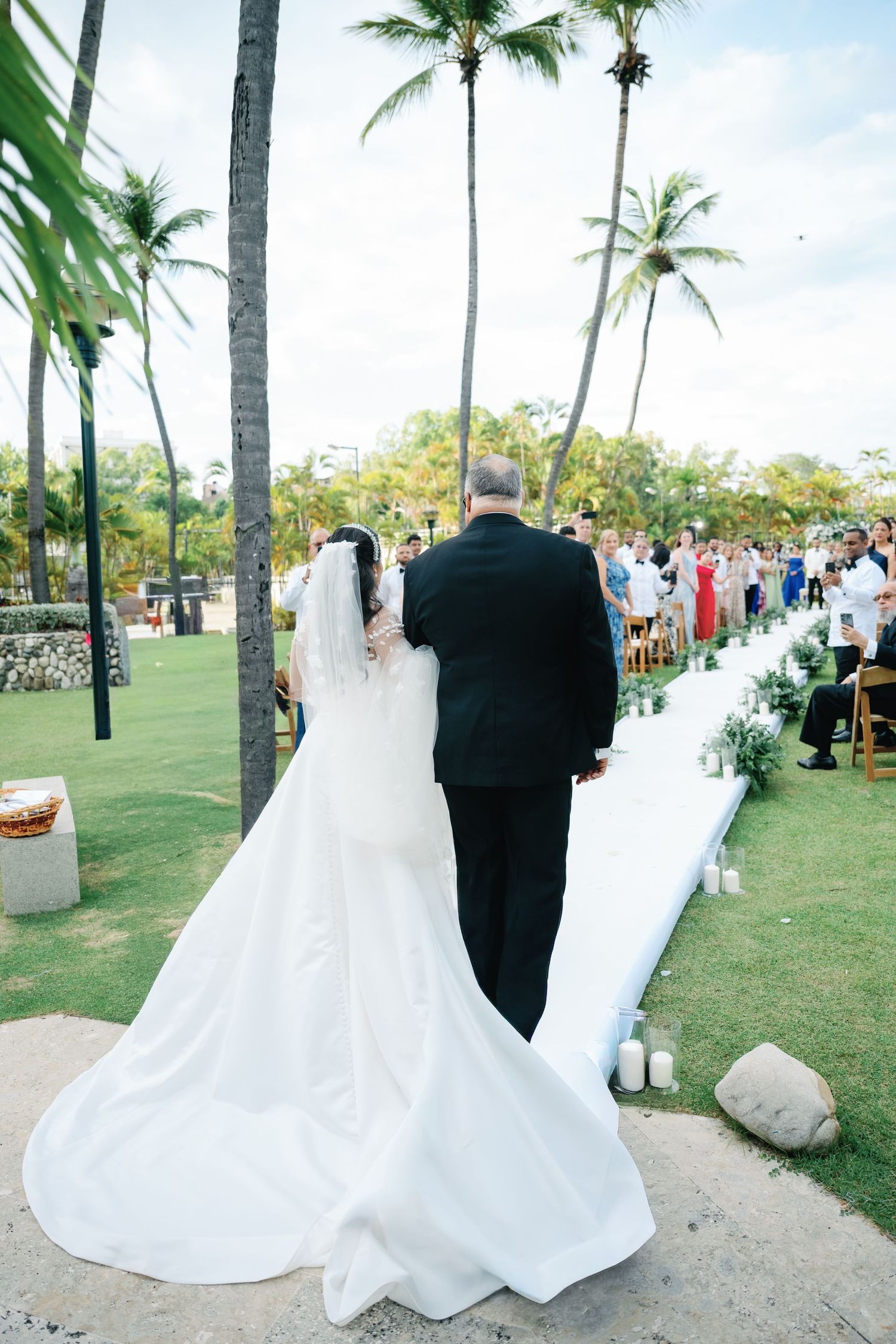 Wedding ceremony on a tropical lawn with palm trees and guests watching a bride and groom walking down a white aisle.