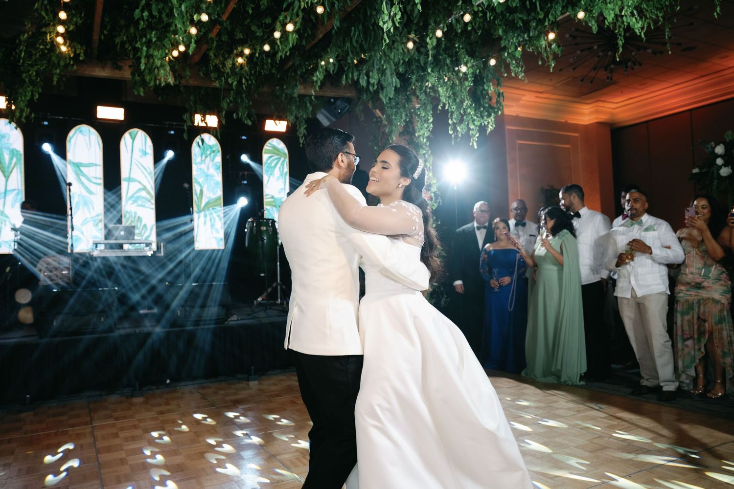 A couple shares a romantic dance on their wedding day as guests watch from the sidelines in a beautifully lit venue.