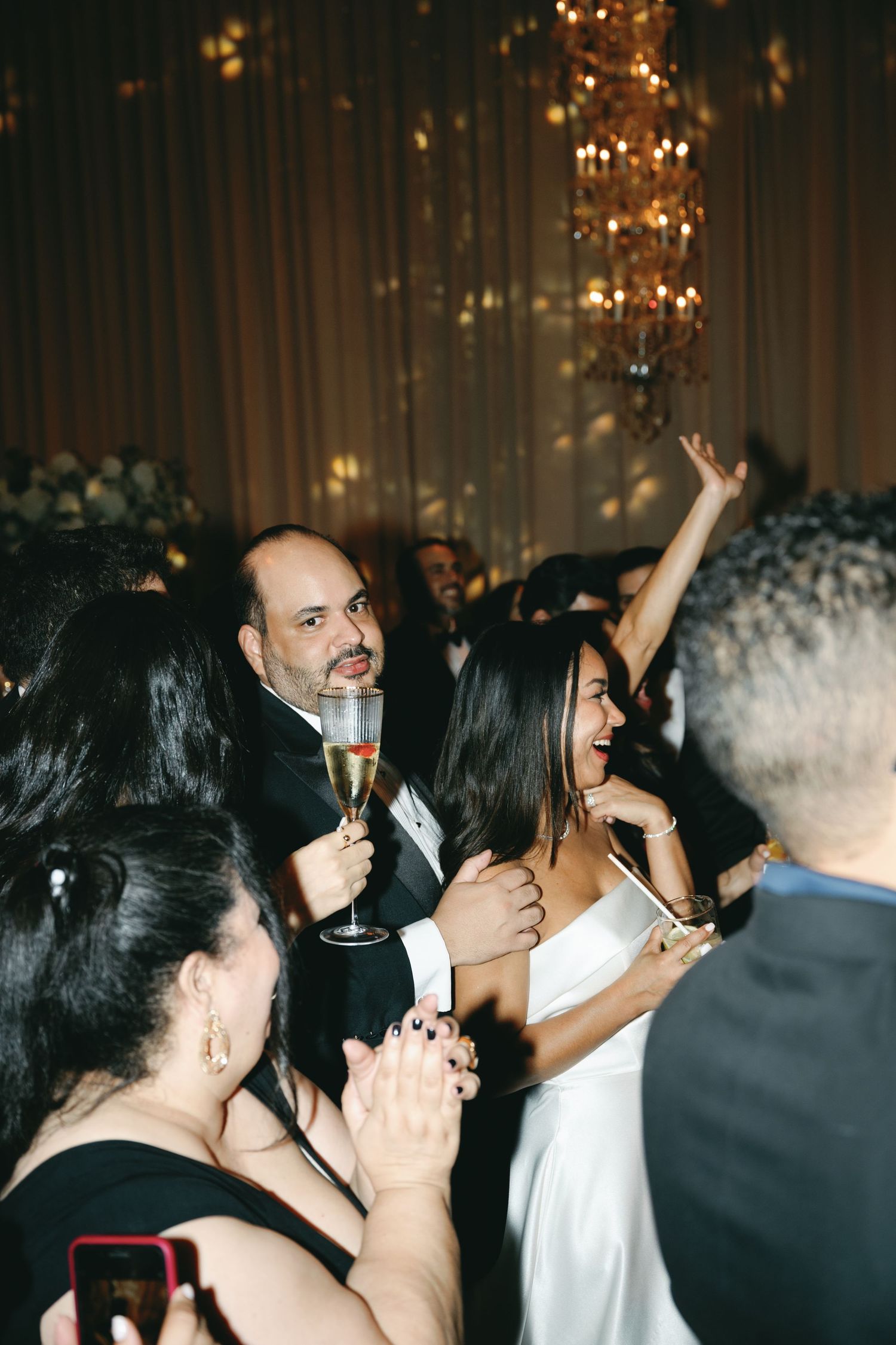 Party guests enjoying drinks and dancing at an elegant event with chandeliers and curtains in the background.