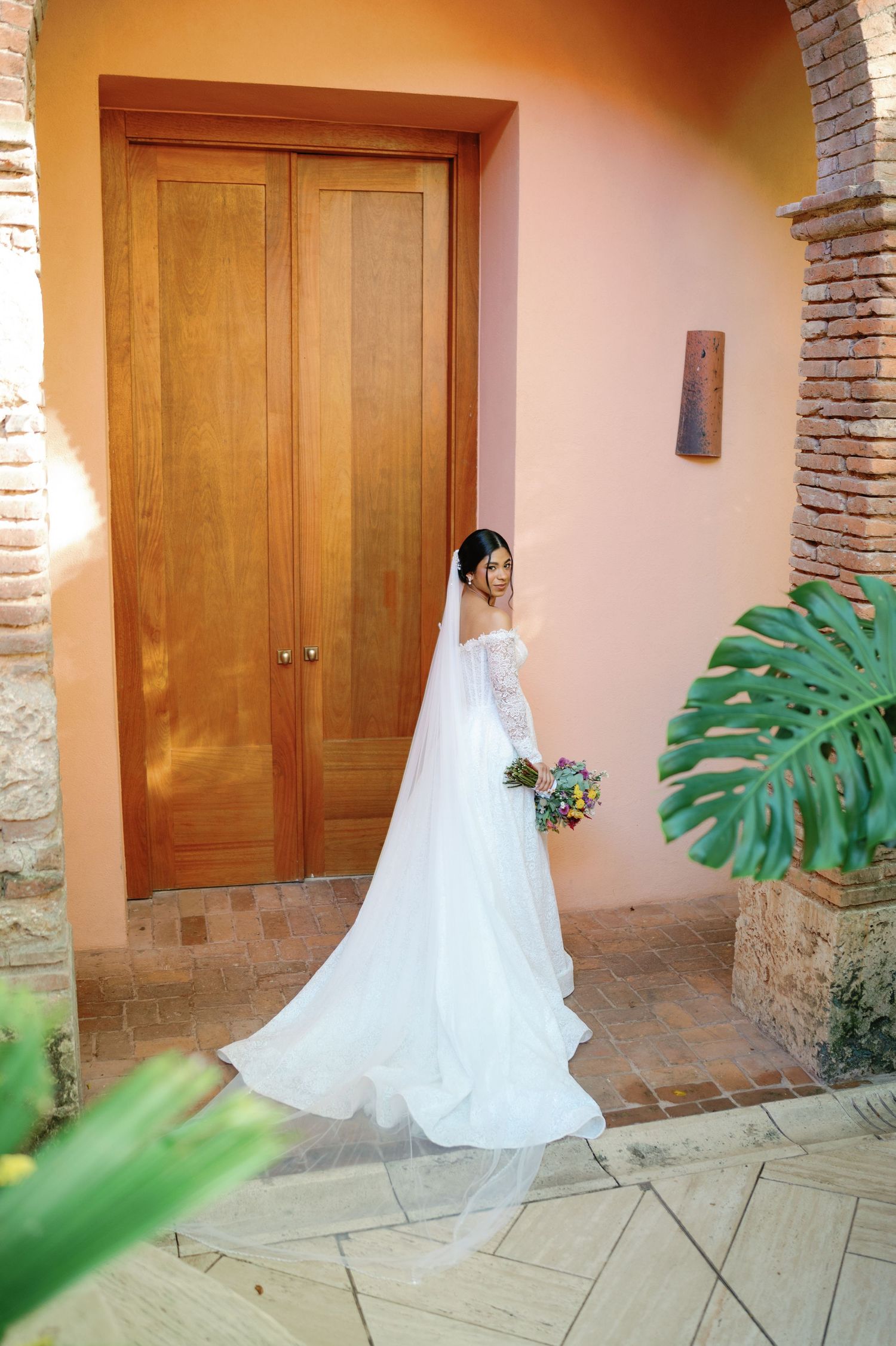 A flowing white wedding dress train trails across stone pavement next to wooden doors and tropical greenery.
