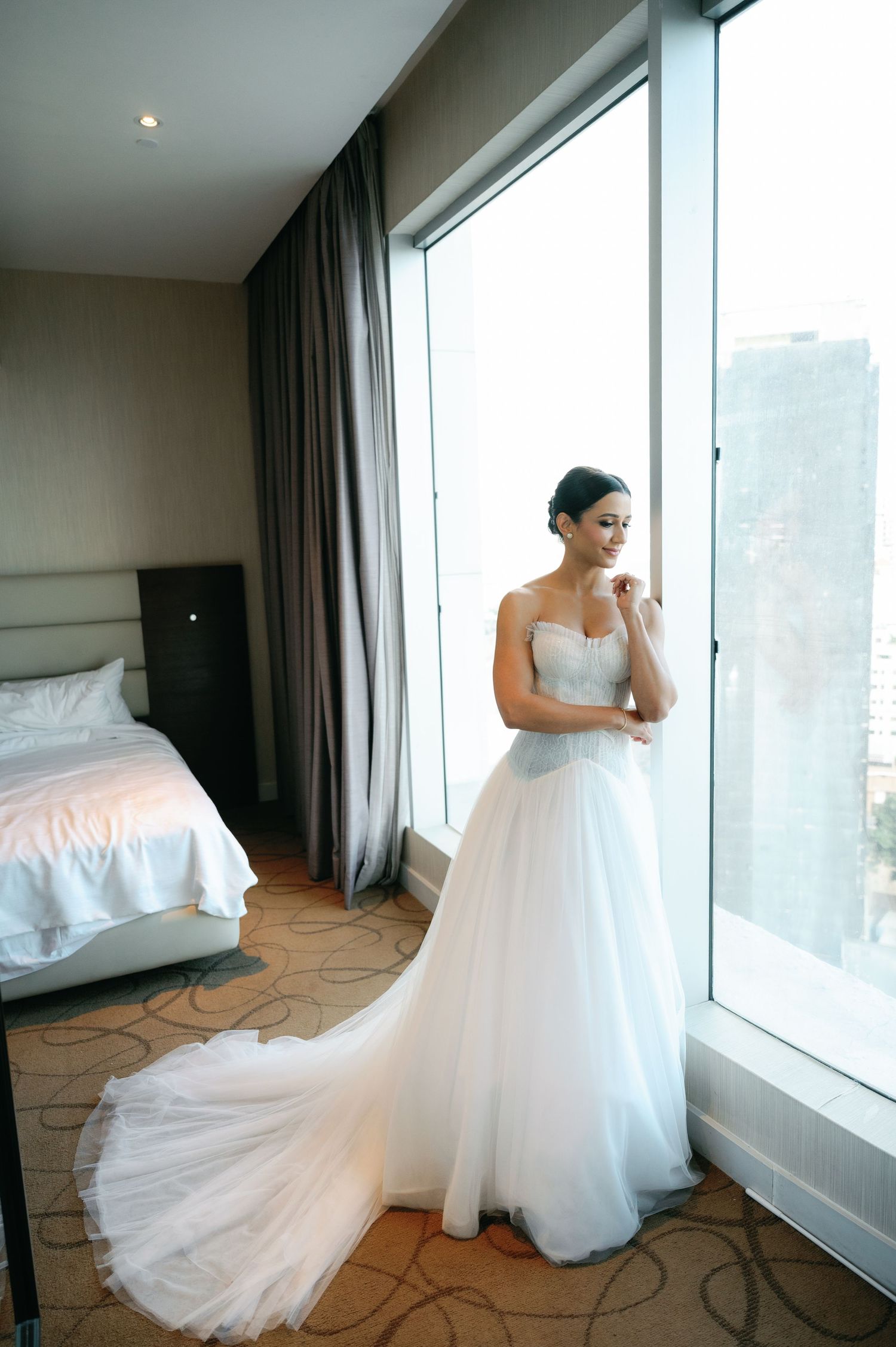 A bride in a flowing white tulle gown poses gracefully by large windows in a modern hotel room.