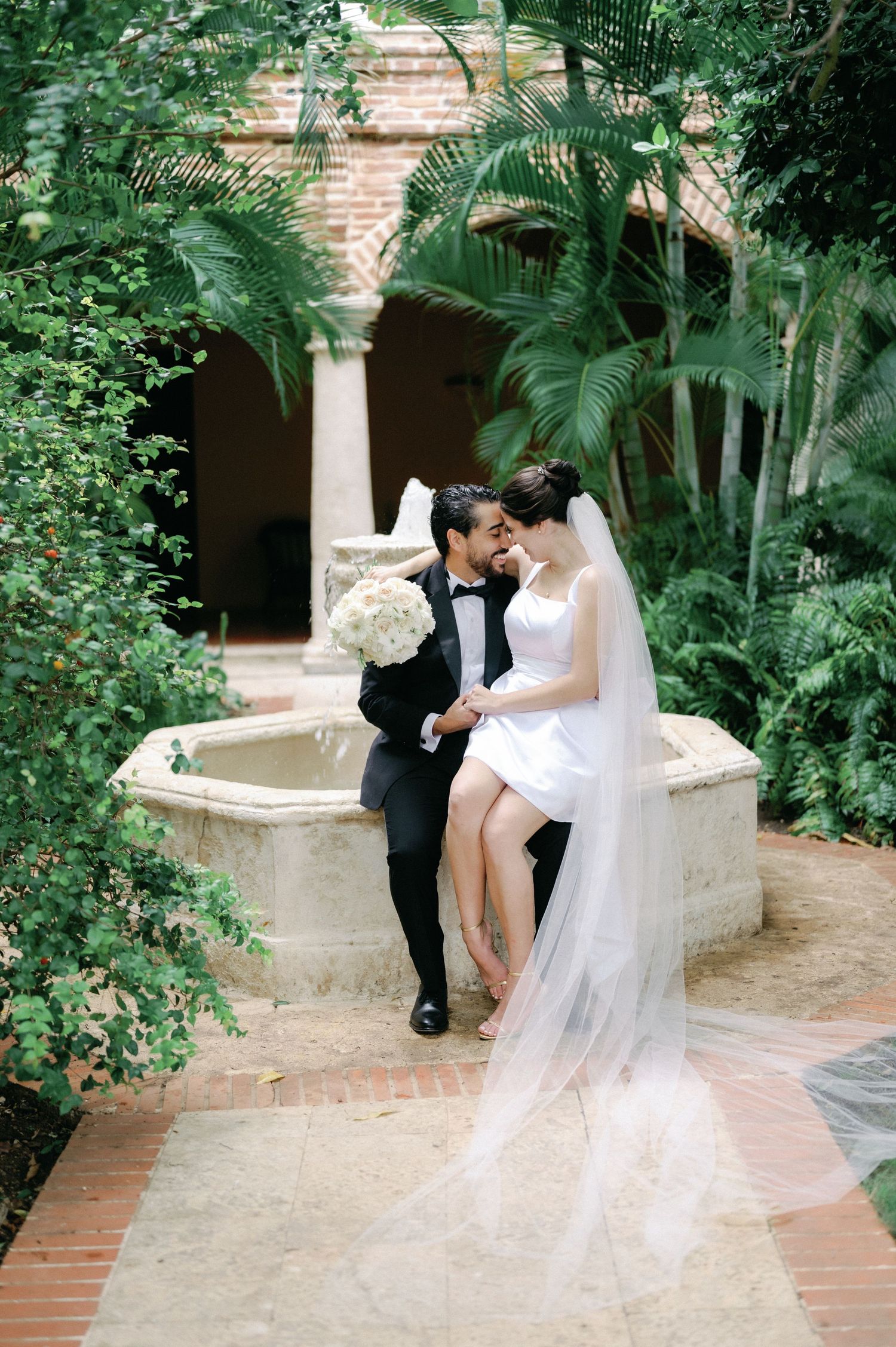 A romantic wedding photo series captured in a lush garden setting with brick columns and greenery.