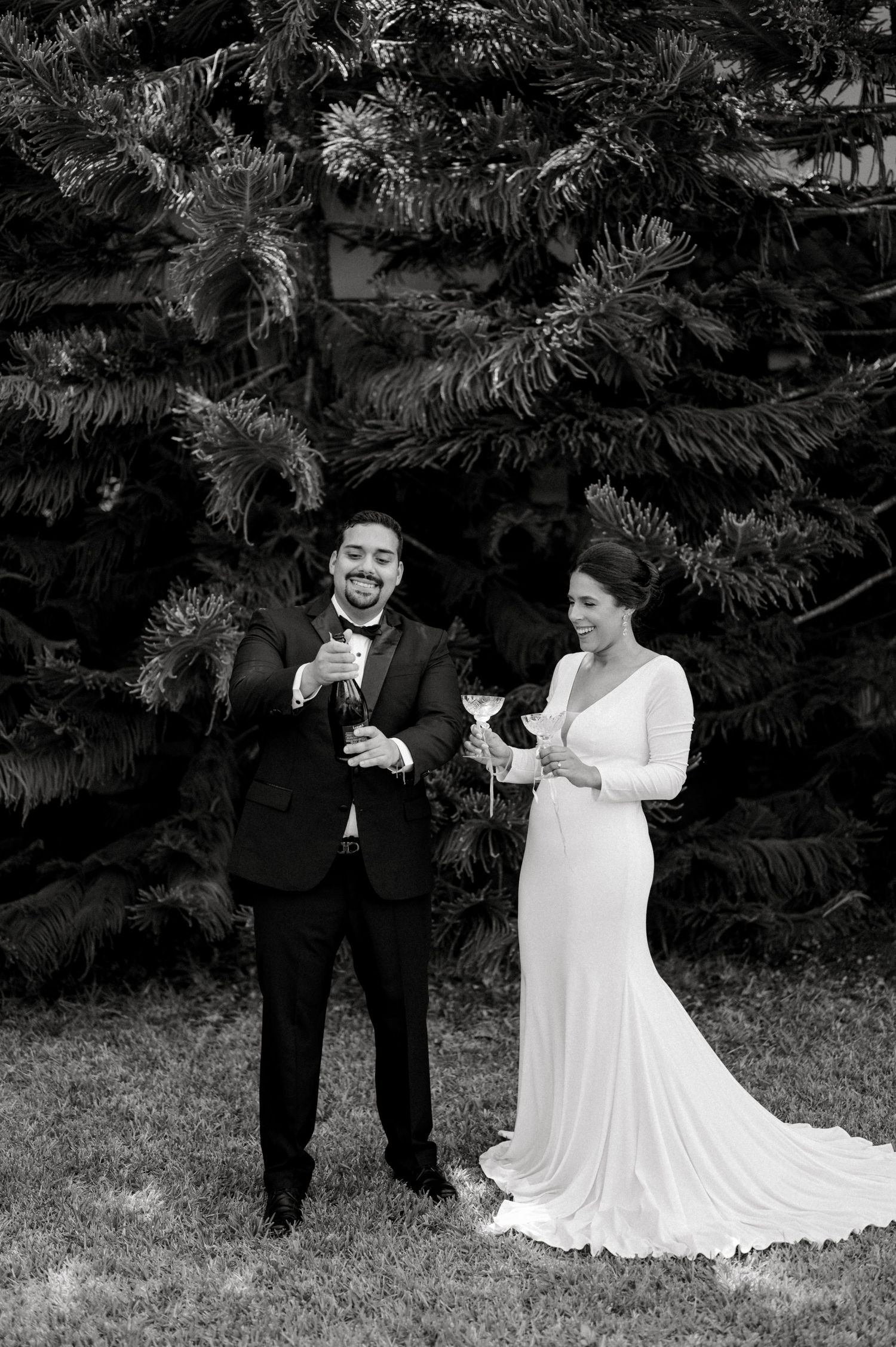A black and white wedding photo showing a couple in formal attire standing in front of evergreen trees.