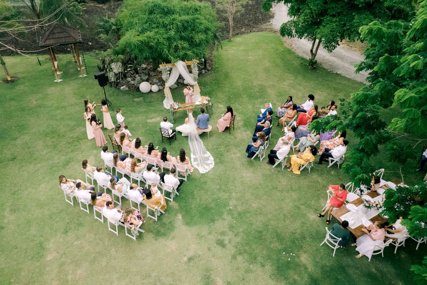 An outdoor garden party with multiple round tables and chairs set up on a lush green lawn surrounded by trees.