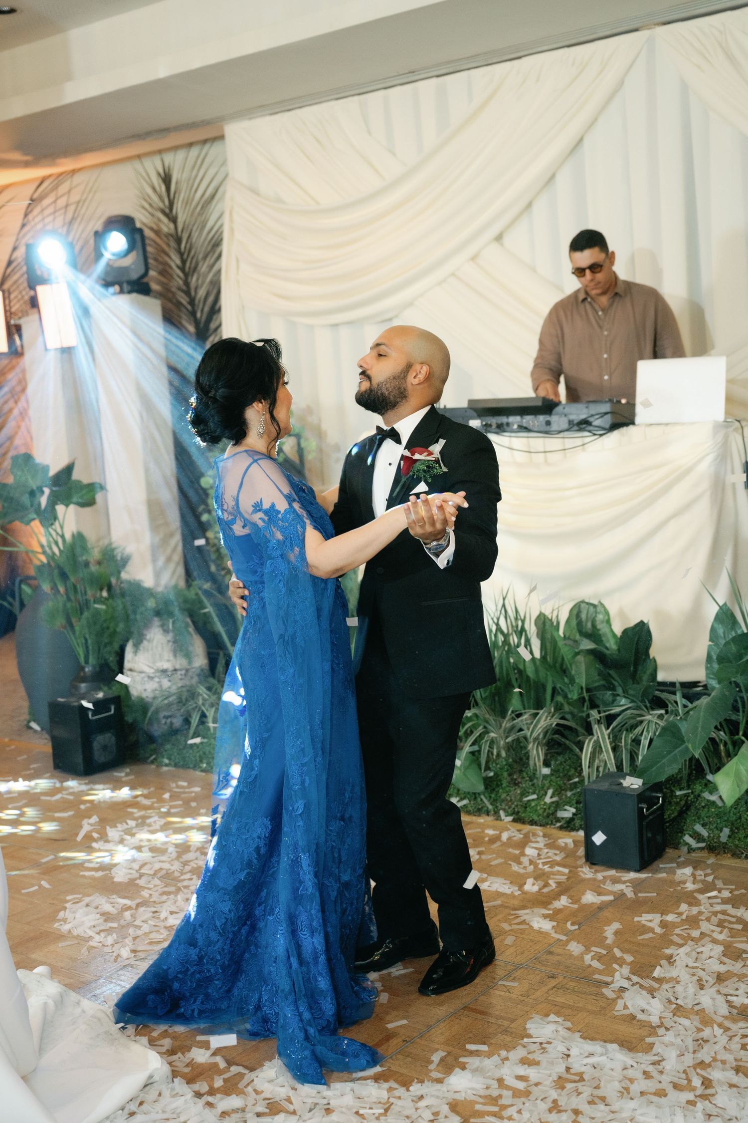 A couple dances together at a wedding reception with white draped walls and scattered petals on the floor.