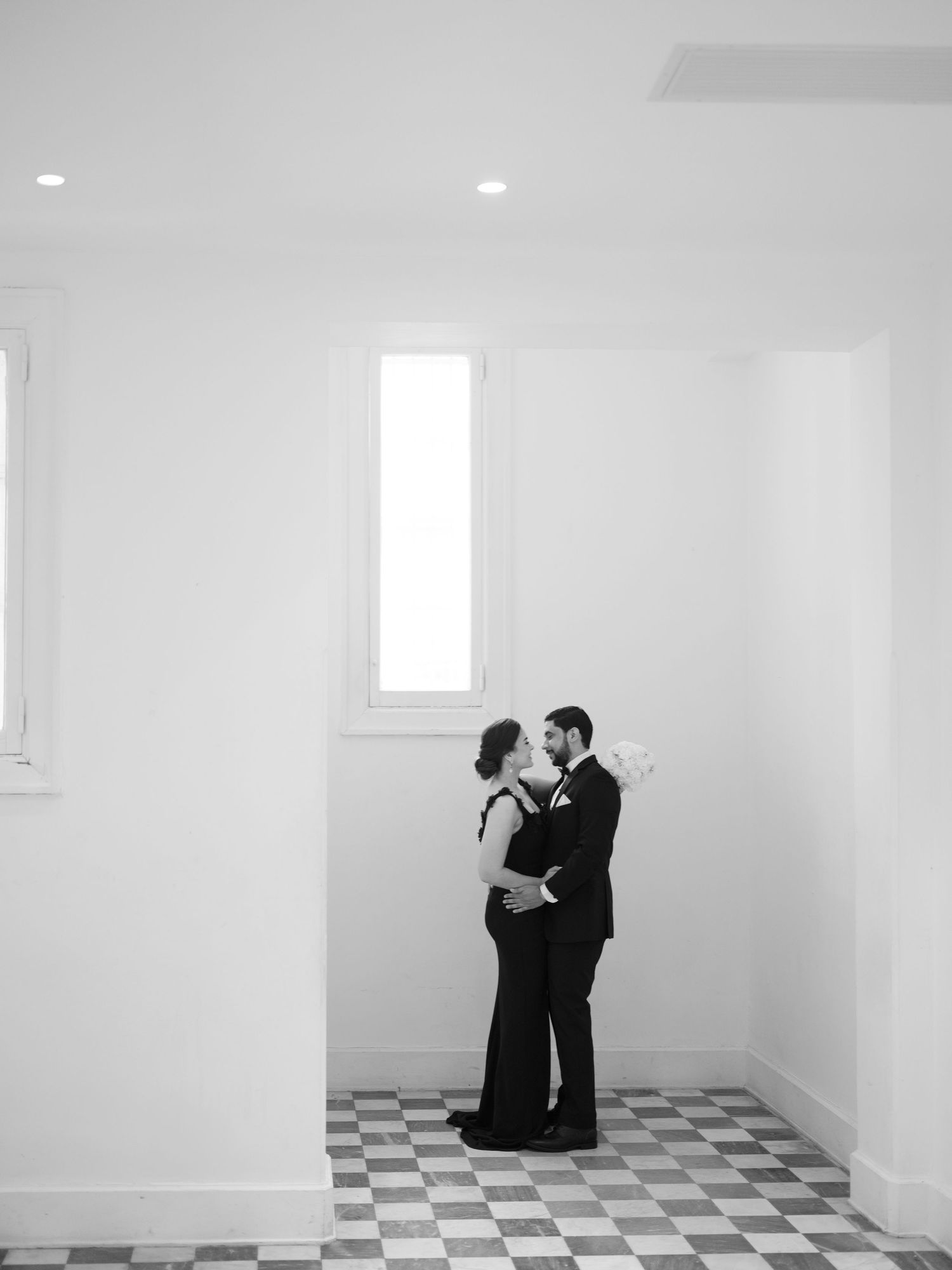Two people in formal black attire dance together in a minimalist white room with checkered floor tiles.