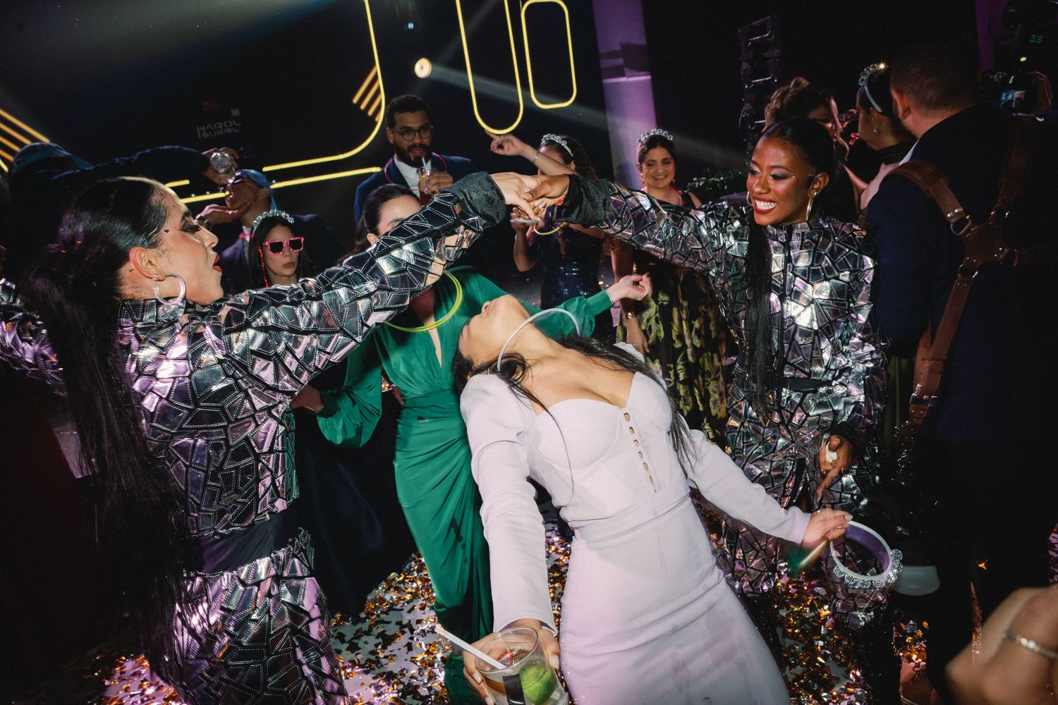 Party crowd dances under colorful lights while performers in metallic costumes entertain on stage.