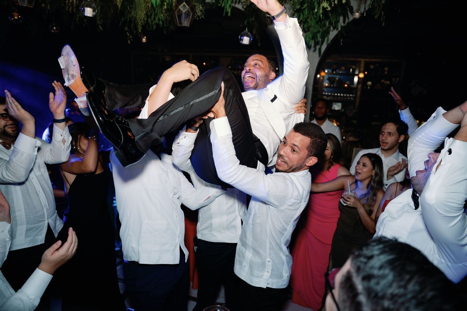 A group of party guests in white dress shirts dance energetically on a dance floor during a celebration.