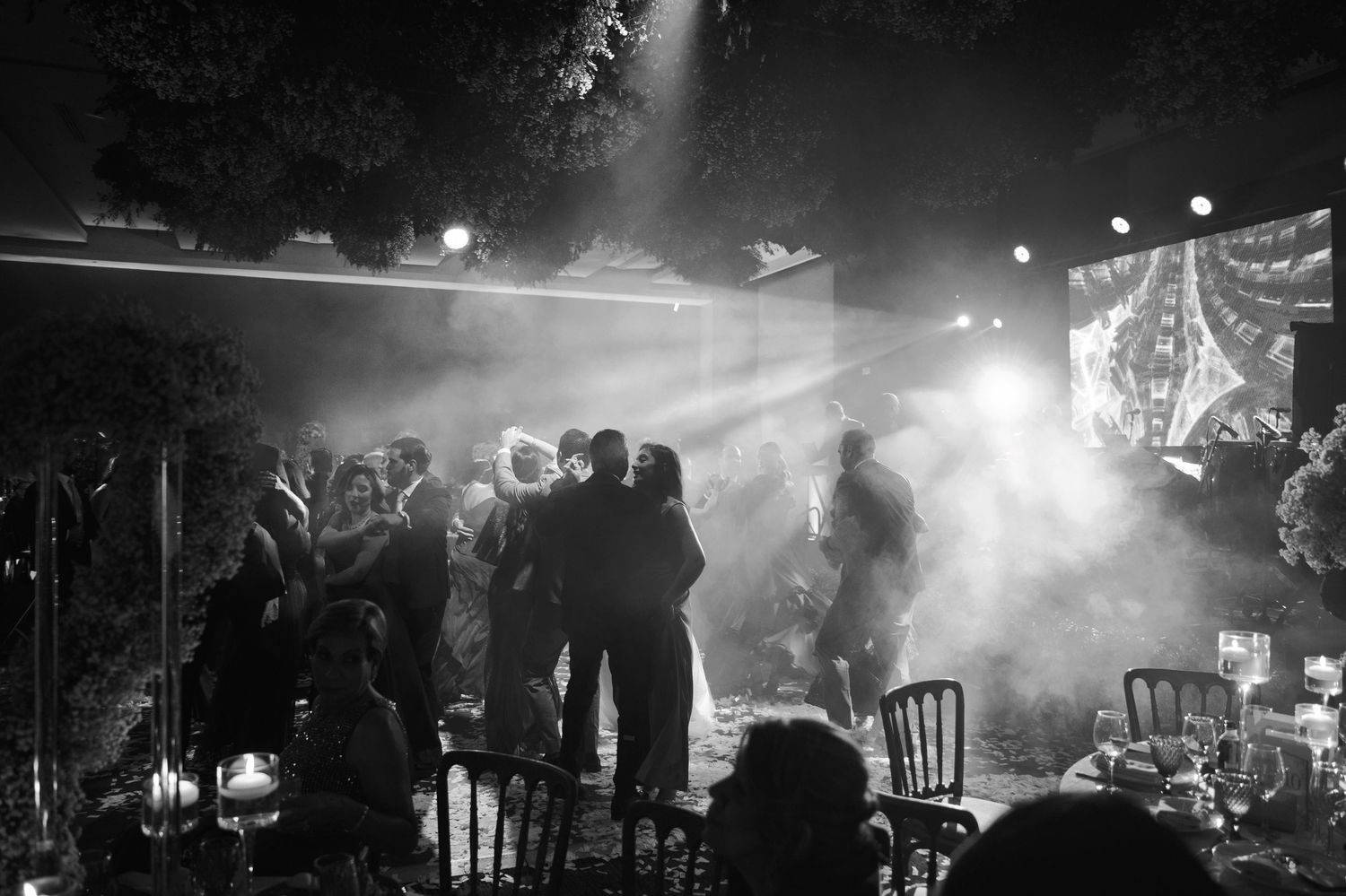 Black and white concert photo showing silhouetted crowd and performers on stage with dramatic light beams and fog effects.
