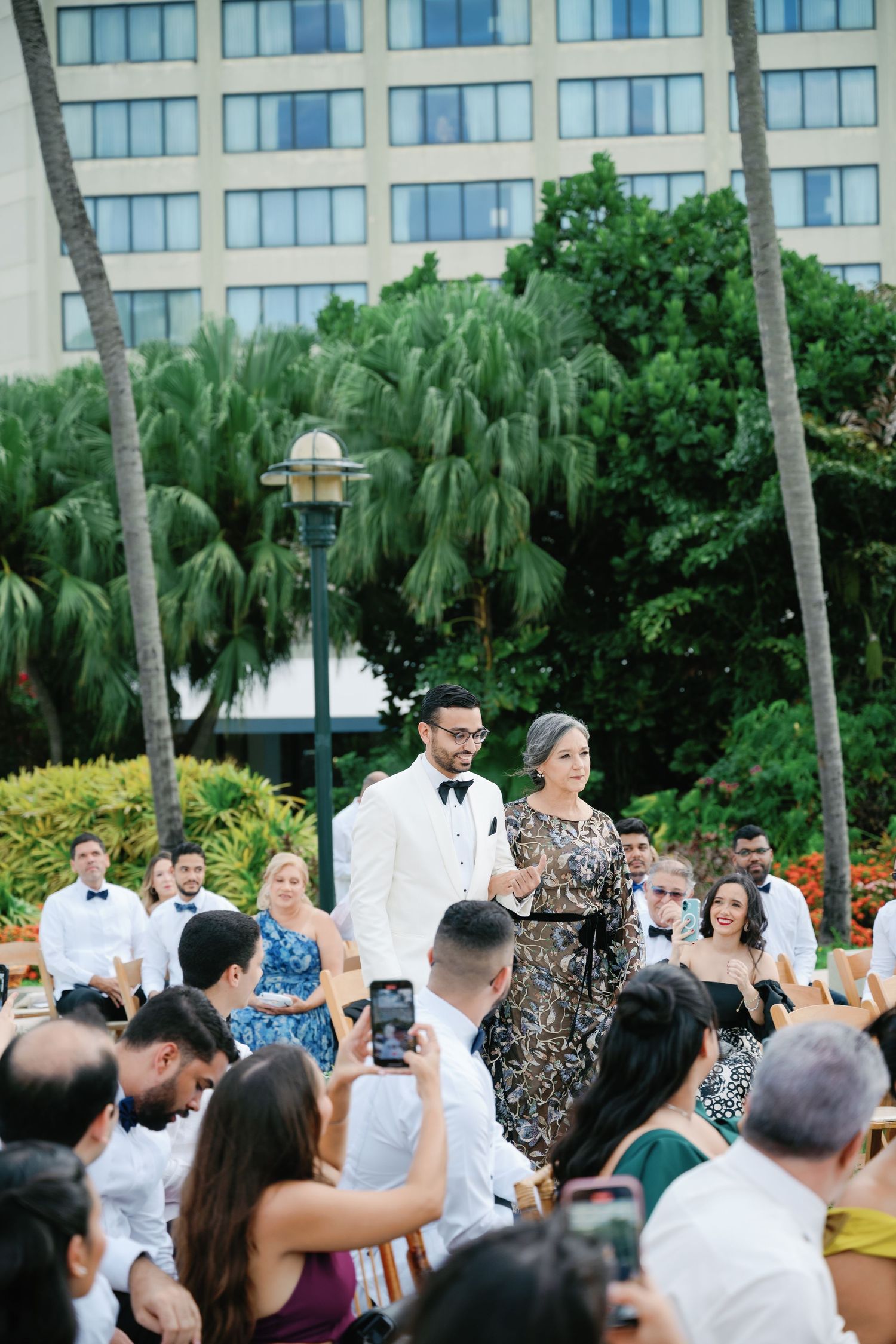 Outdoor wedding reception at a beachfront resort with guests capturing moments on their phones.