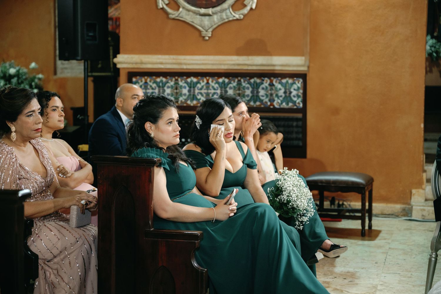Wedding guests react emotionally during a ceremony while seated in emerald green and blush colored attire.