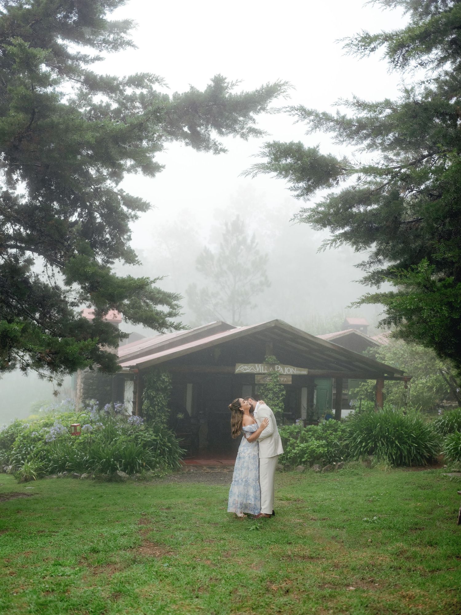 Misty morning scene at a rustic cabin surrounded by evergreen trees and lush grass in a peaceful mountain setting.