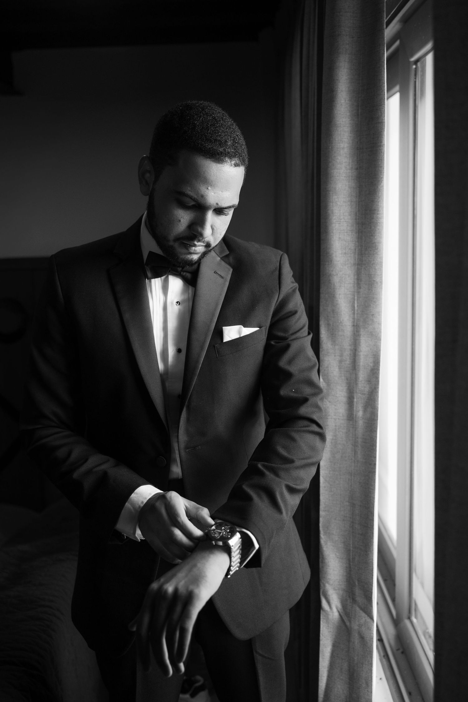 Dramatic black and white portrait showing a groom adjusting his tie near a window.