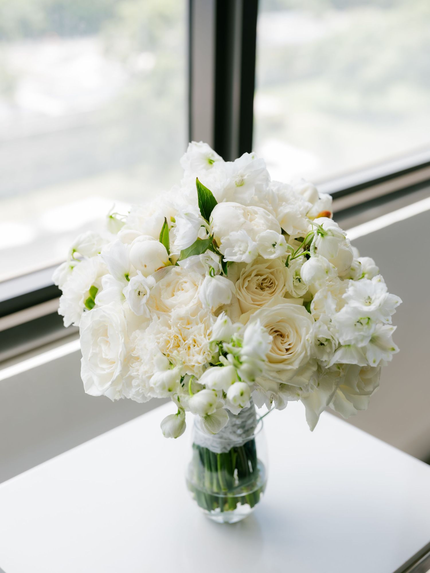 An elegant white wedding bouquet with roses and mixed flowers sits in a glass vase by a window.