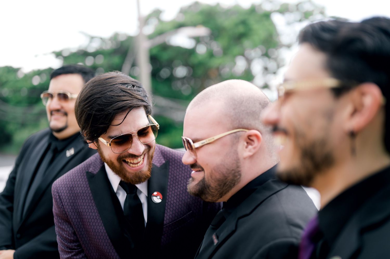 A group of groomsmen share a candid laughing moment together outdoors at a wedding celebration.