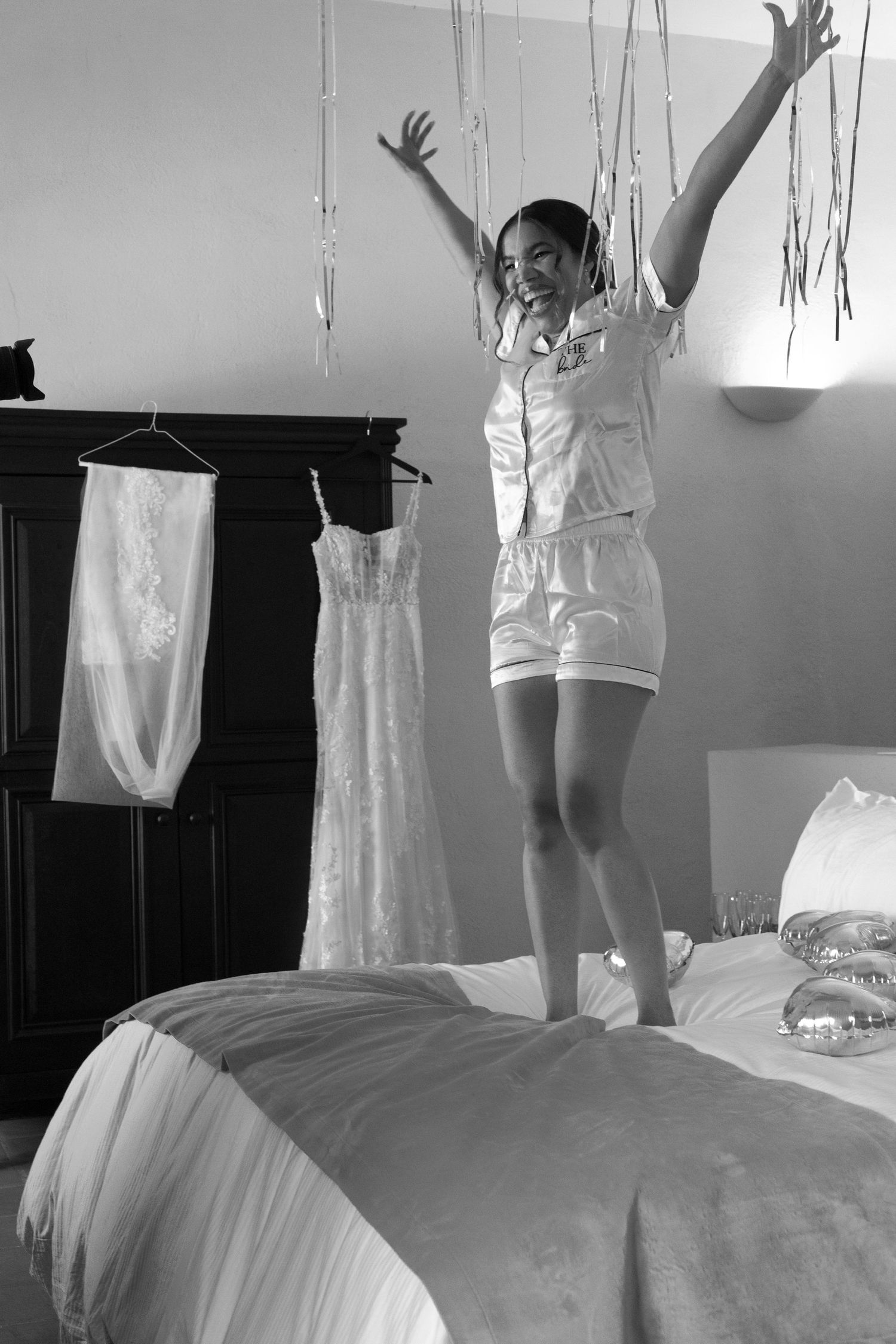A person in white pajamas jumps joyfully on a bed with a wedding dress hanging in the background.