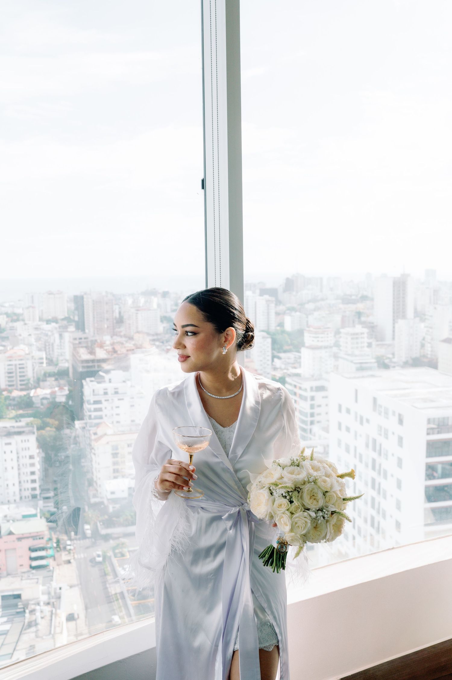 Woman in white robe holds white floral bouquet while looking out window at city skyline view.