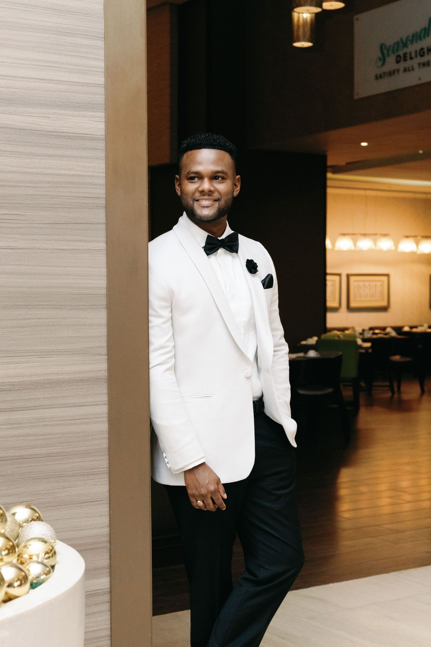 Groom poses confidently in a white tuxedo jacket and black pants in an elegant venue hallway.