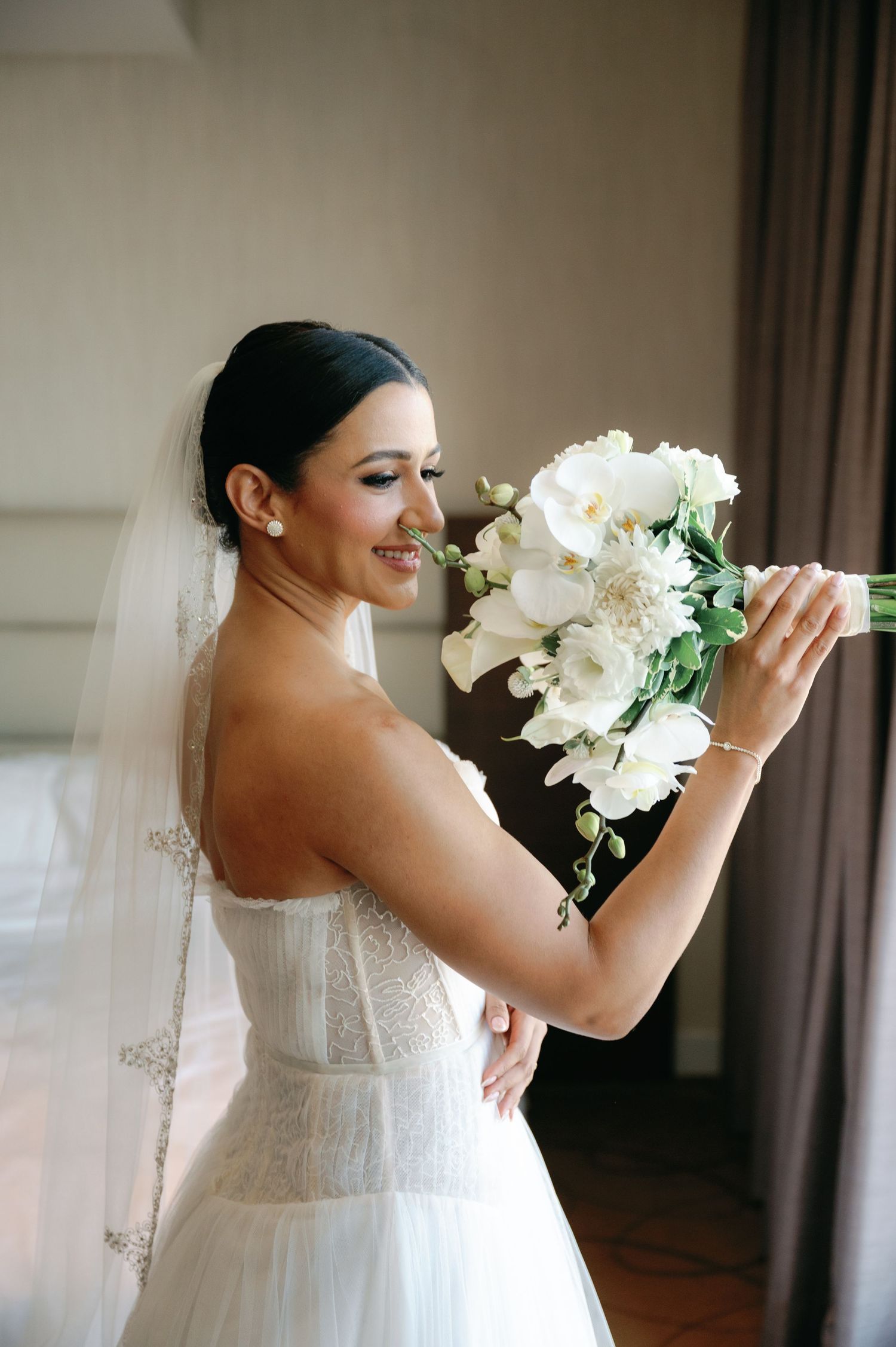 A bride in a white wedding dress holds and smells a bouquet of white flowers while smiling joyfully.