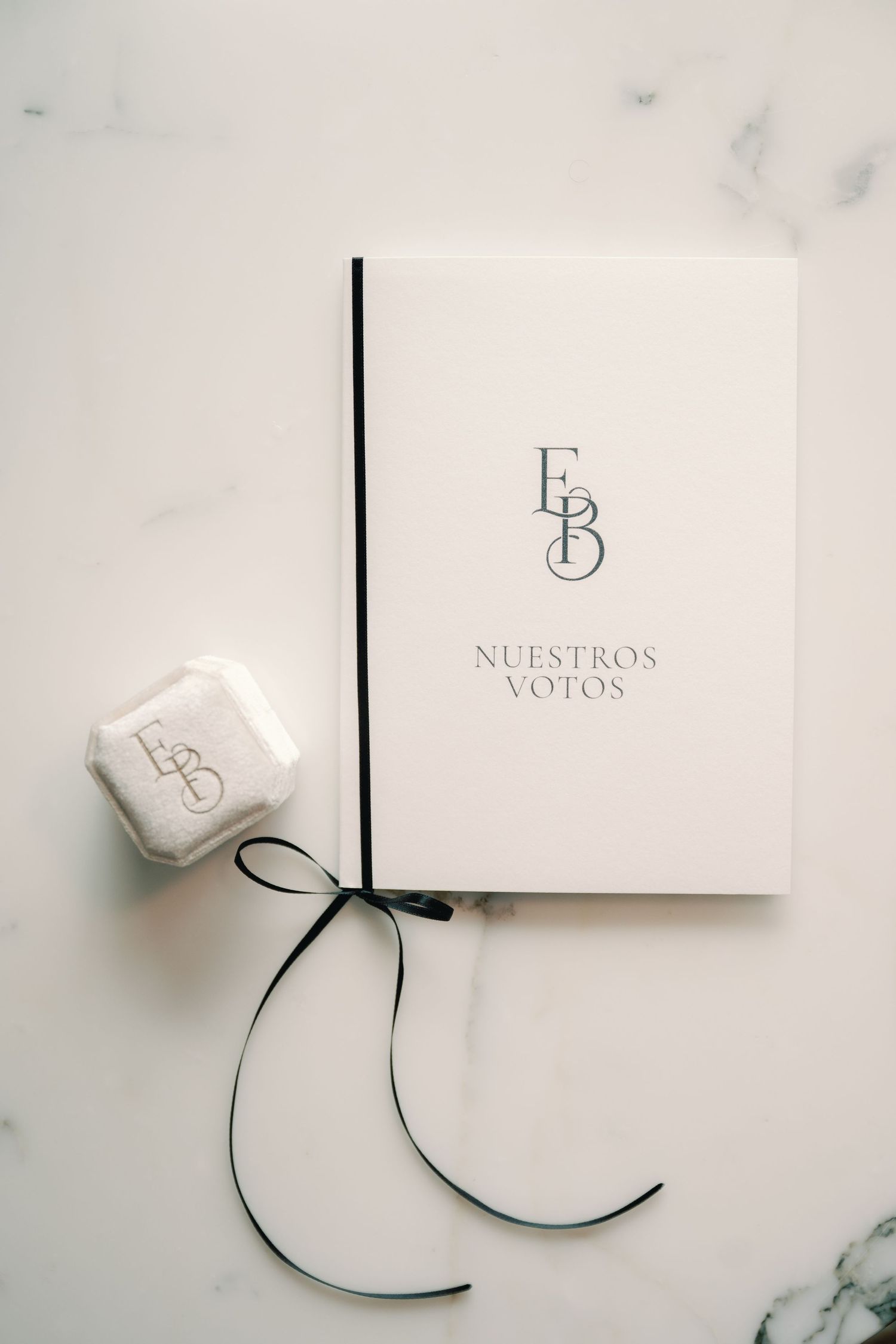 Elegant white wedding vow book with monogram and black ribbon on marble surface next to small ring box.