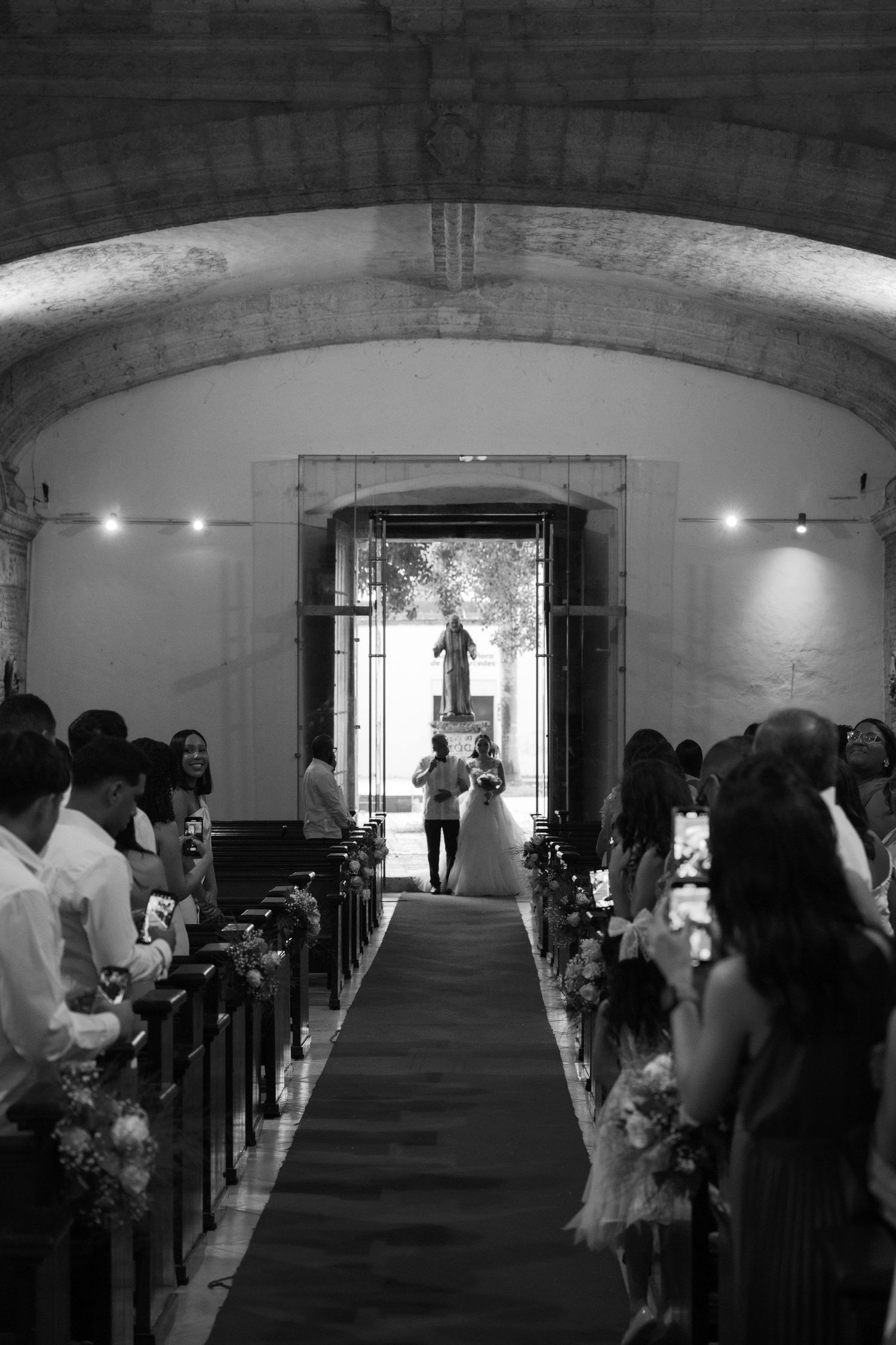 A black and white wedding ceremony photo showing guests in church pews with a carpeted aisle and arched doorway.