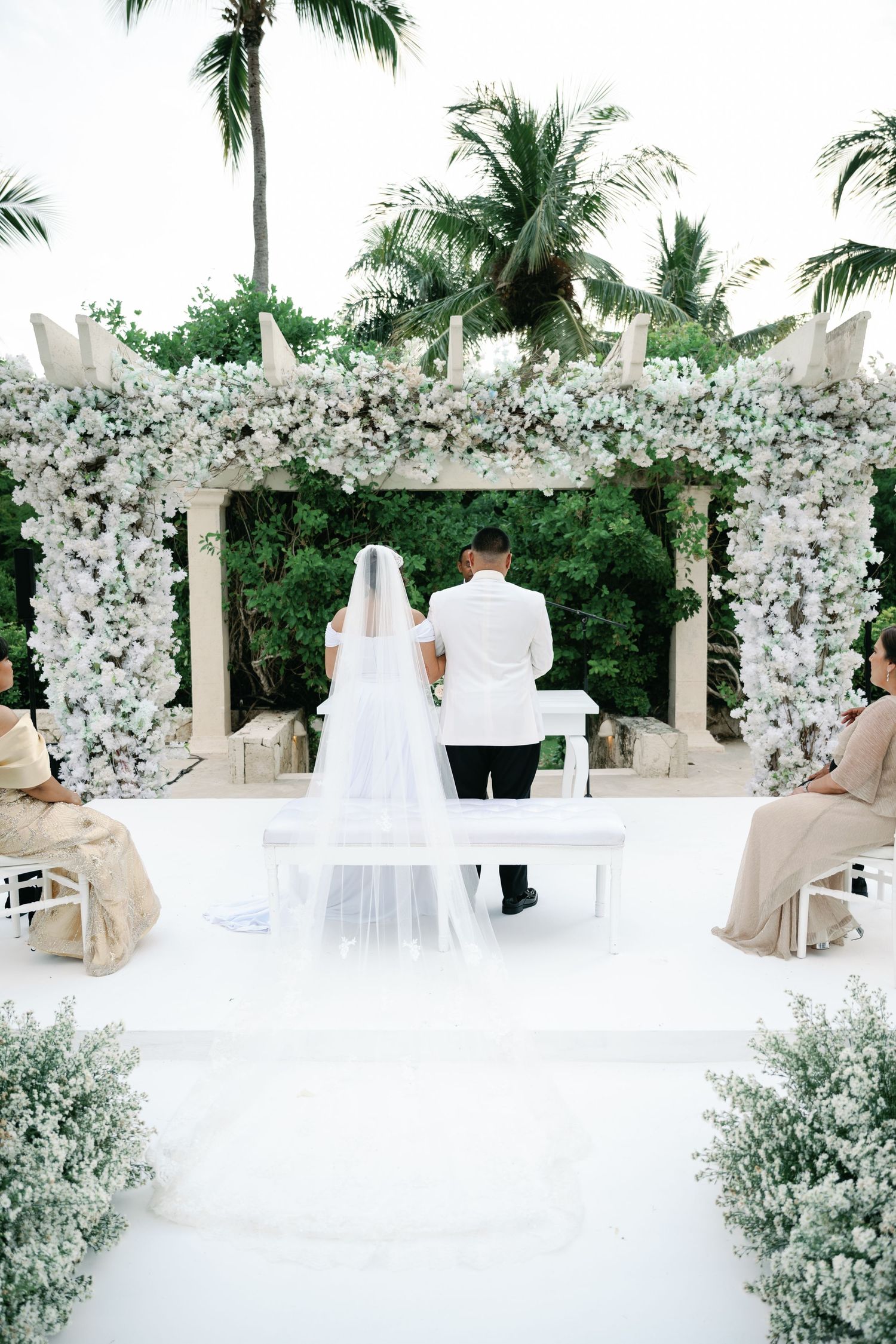 A wedding ceremony setup with white floral arch, palm trees, and elegant white decor in a tropical outdoor setting.