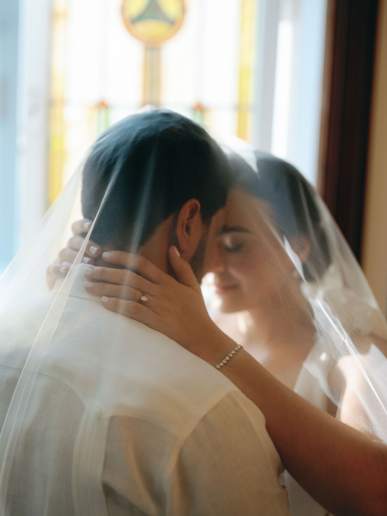 A tender moment between newlyweds under a delicate white veil, with soft light streaming through windows in the background.