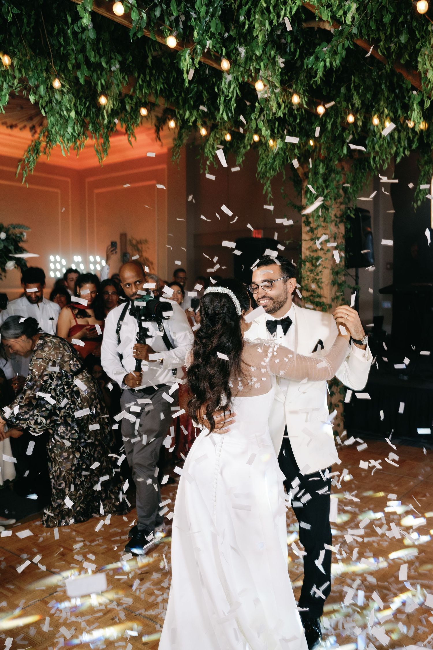 Wedding celebration with guests throwing paper airplanes in an elegant venue decorated with hanging greenery and lights.