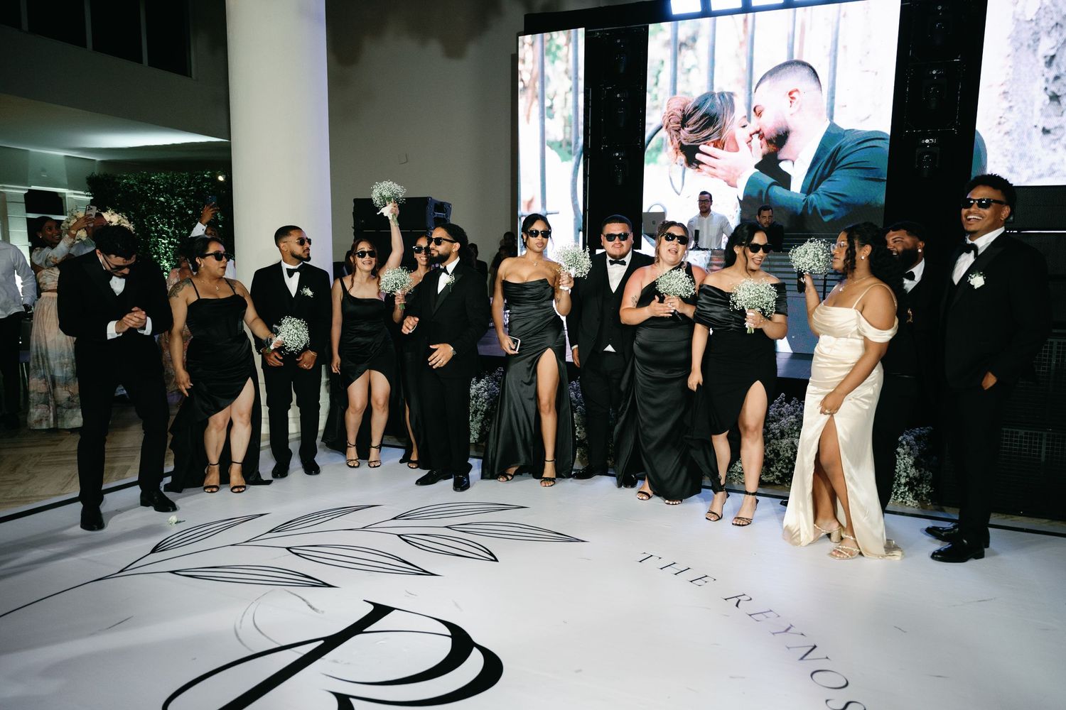 Wedding party in black formal attire poses on a dance floor with decorative monogram design.