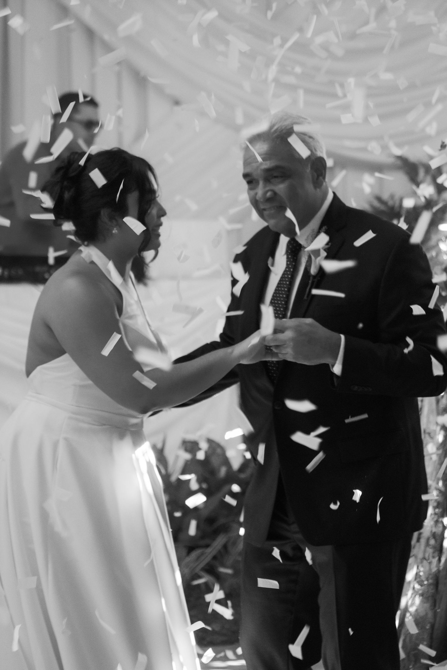 A couple shares a joyful dance moment at their wedding celebration as confetti falls around them in black and white.