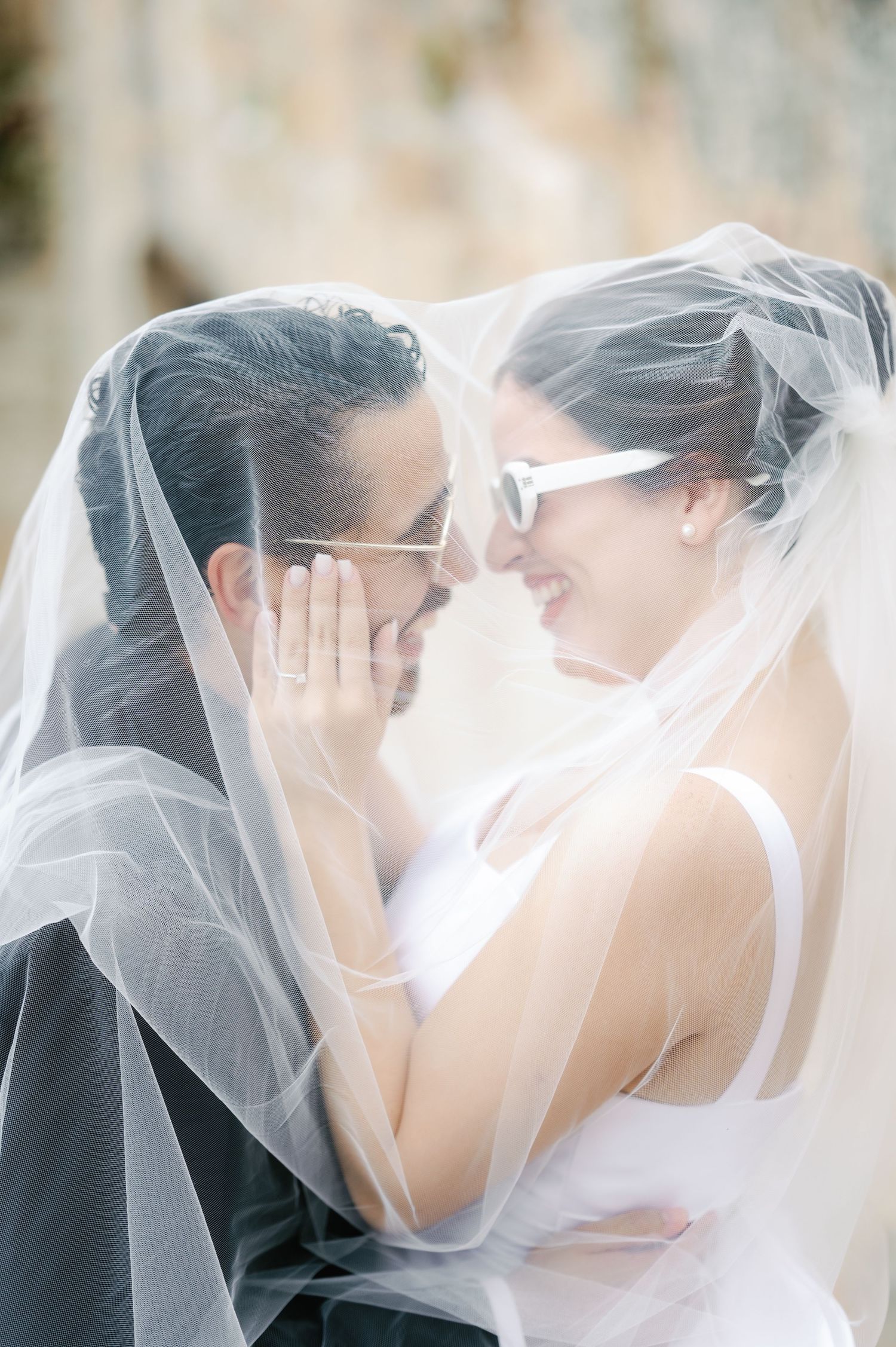 A romantic wedding couple embraces under a flowing white veil in soft lighting.