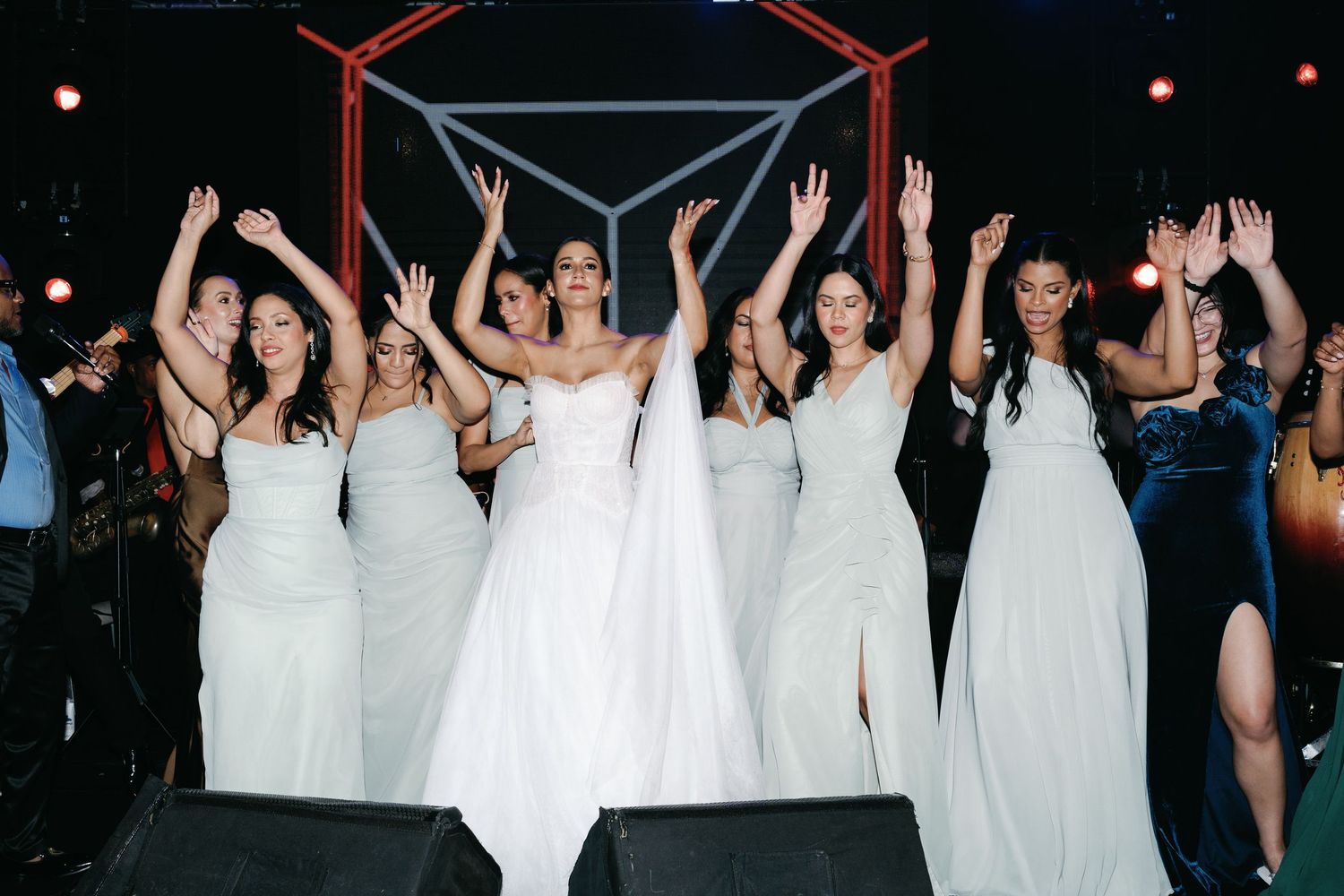 A group of bridesmaids in white dresses raise their arms in celebration during a wedding reception.