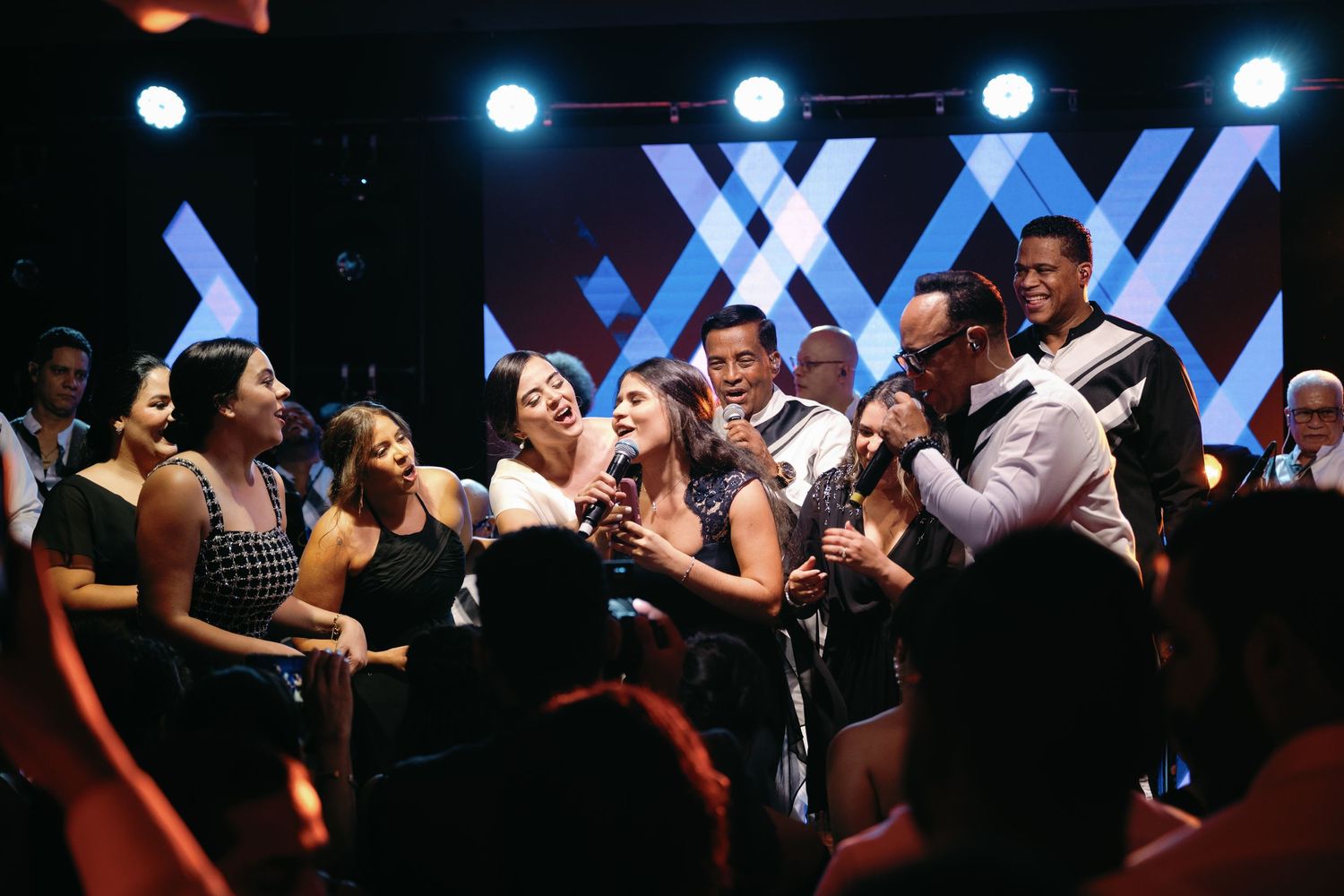Performers on stage during a concert with blue lighting and an enthusiastic crowd in the background.