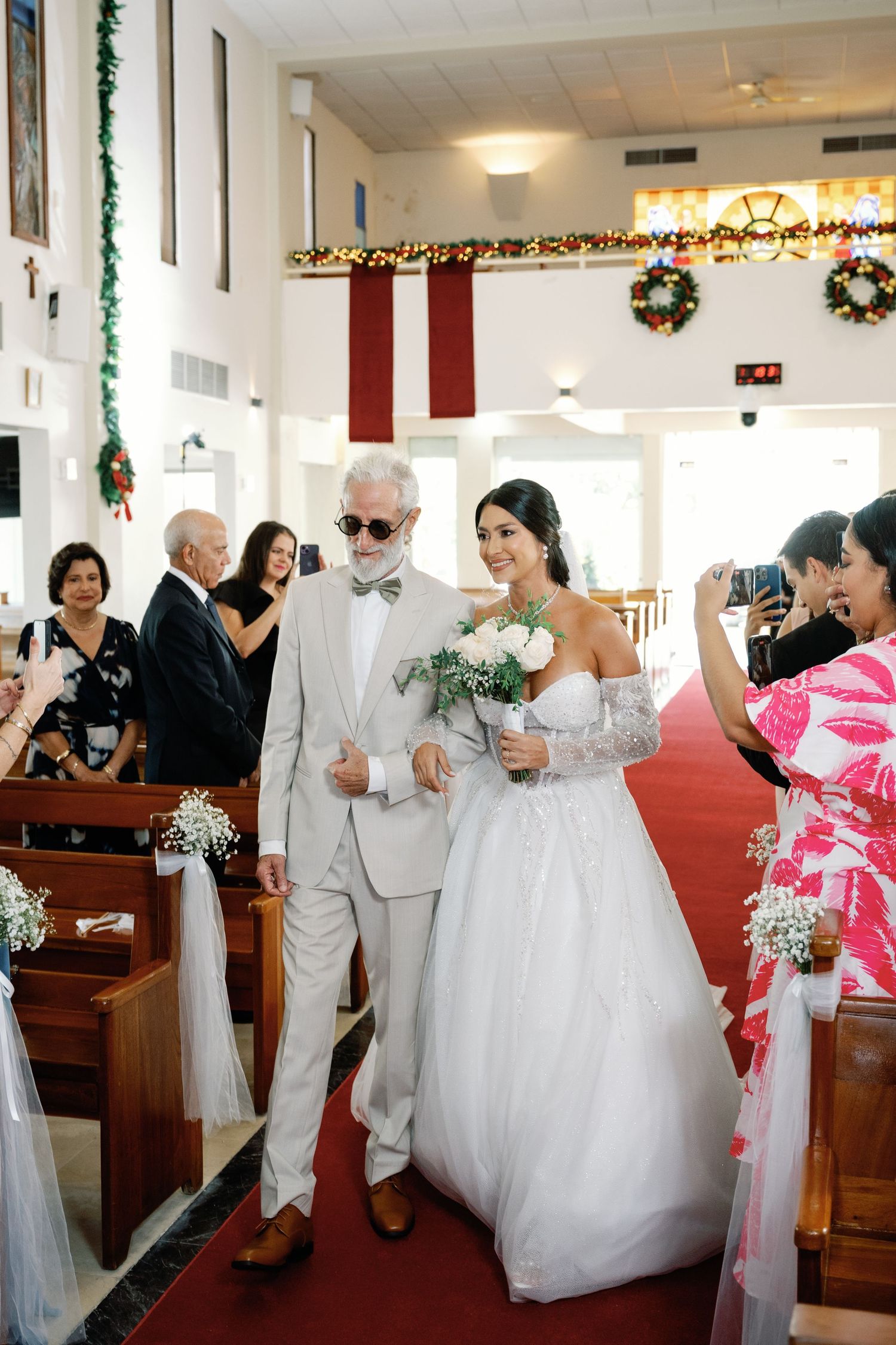 A bride in a white gown walks down the church aisle with her escort during a wedding ceremony decorated with wreaths.