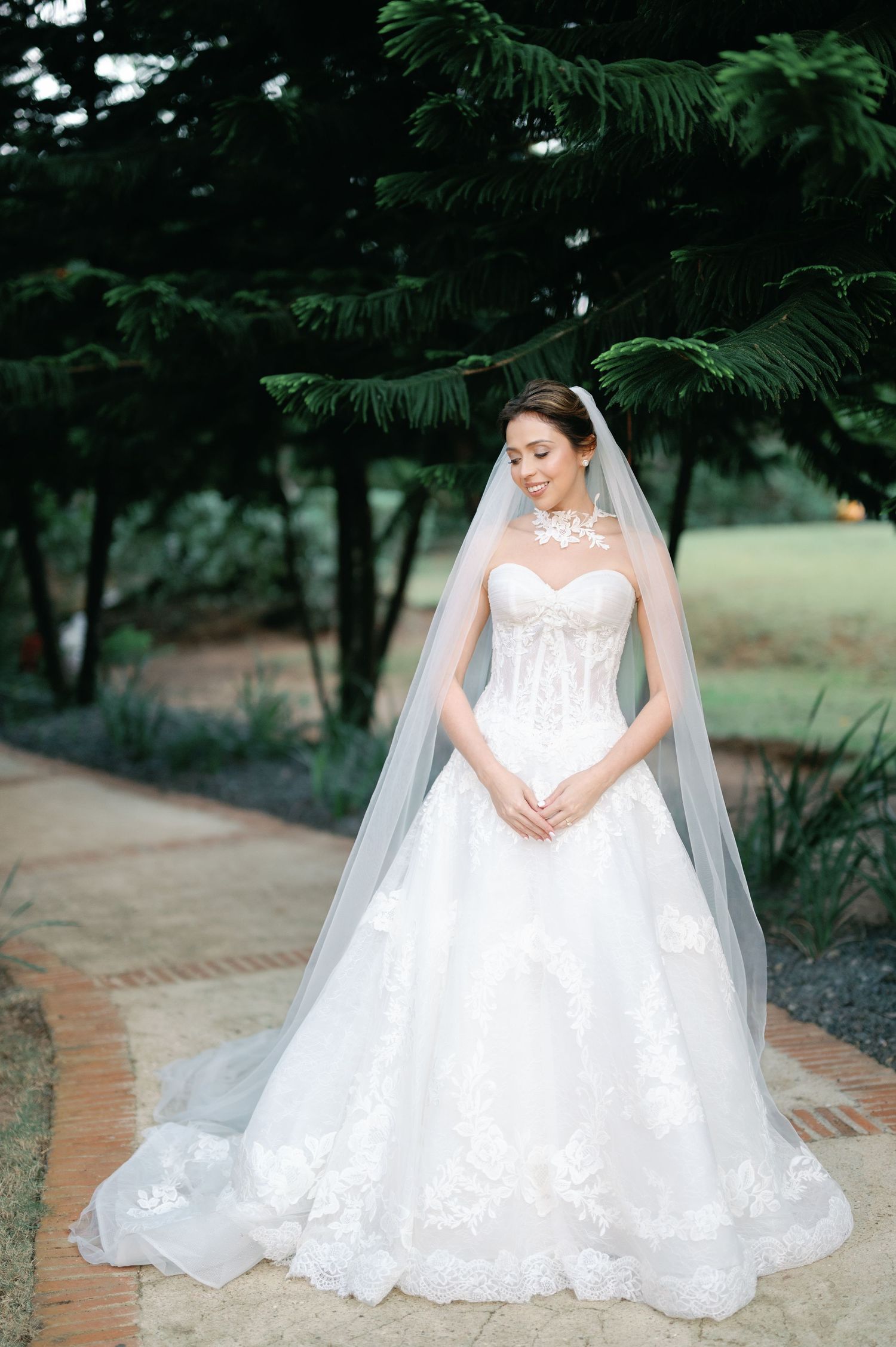 A bride in a flowing white lace wedding gown sits gracefully on stone steps surrounded by lush greenery.