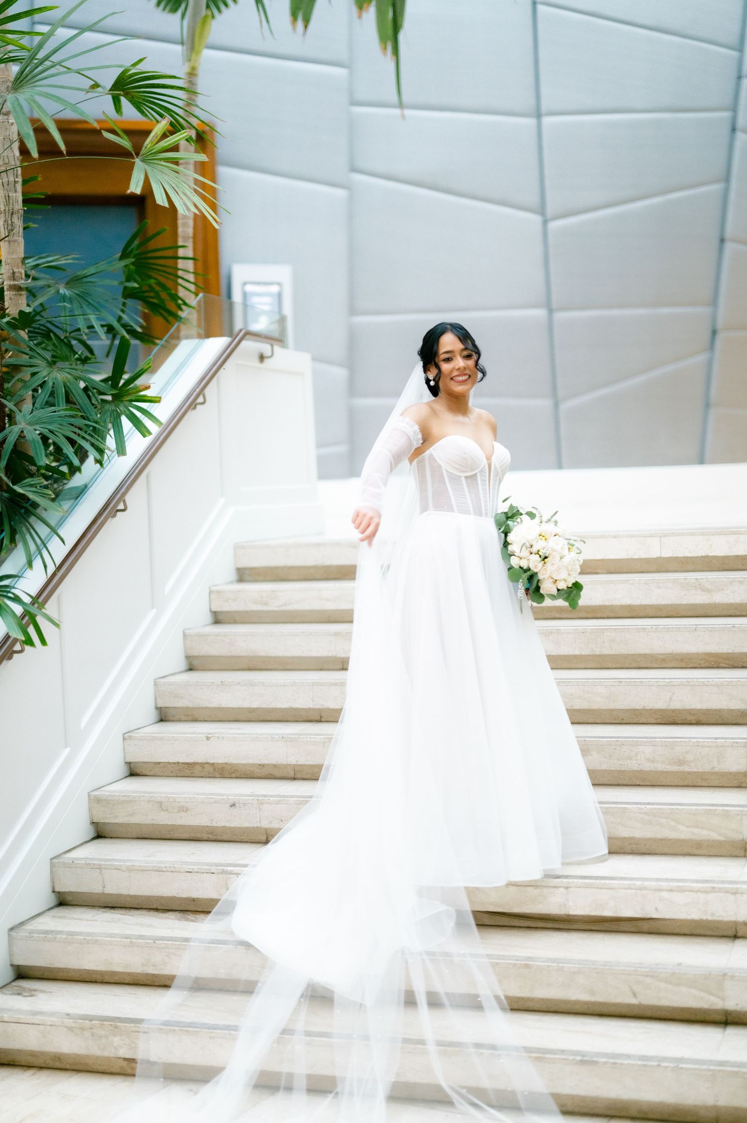 A bride in a flowing white wedding gown with long veil stands gracefully on marble stairs near tropical plants.