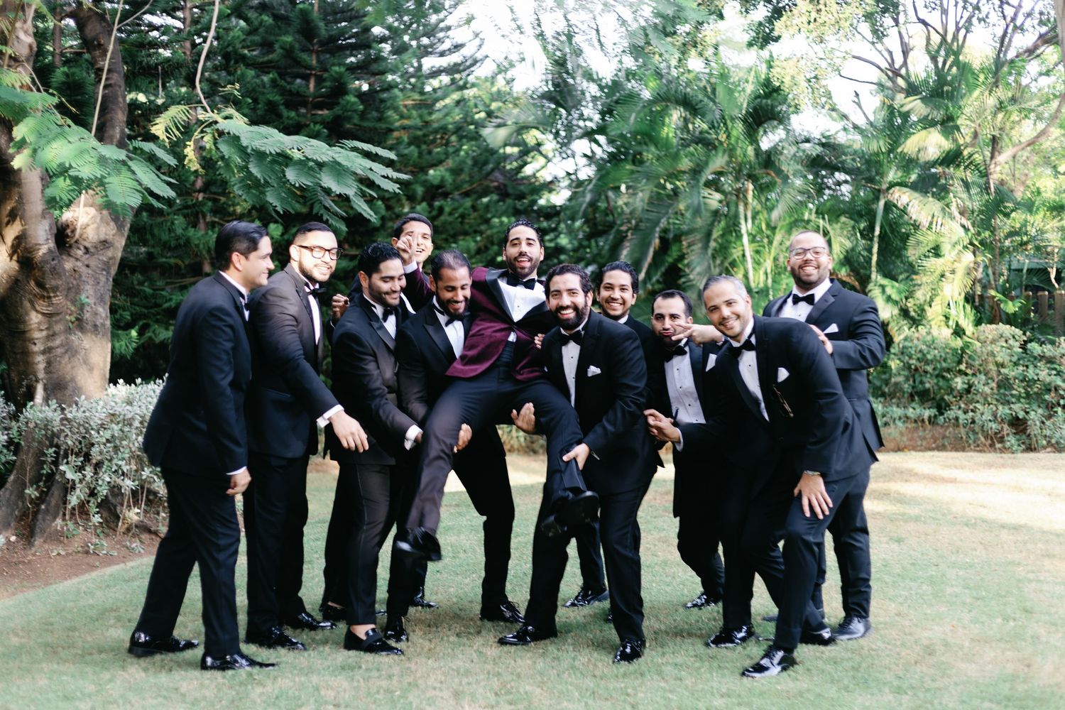 Wedding party in dark suits poses playfully in a garden setting during outdoor photo session.