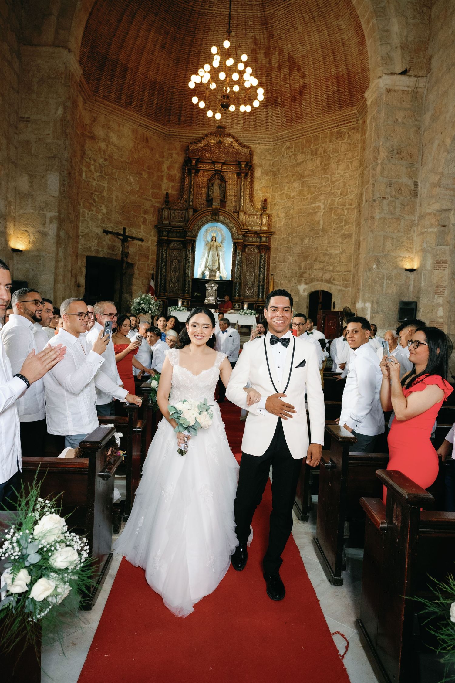 Wedding party celebrates with joyful dancing and raised hands inside a rustic stone church venue with vaulted ceilings.