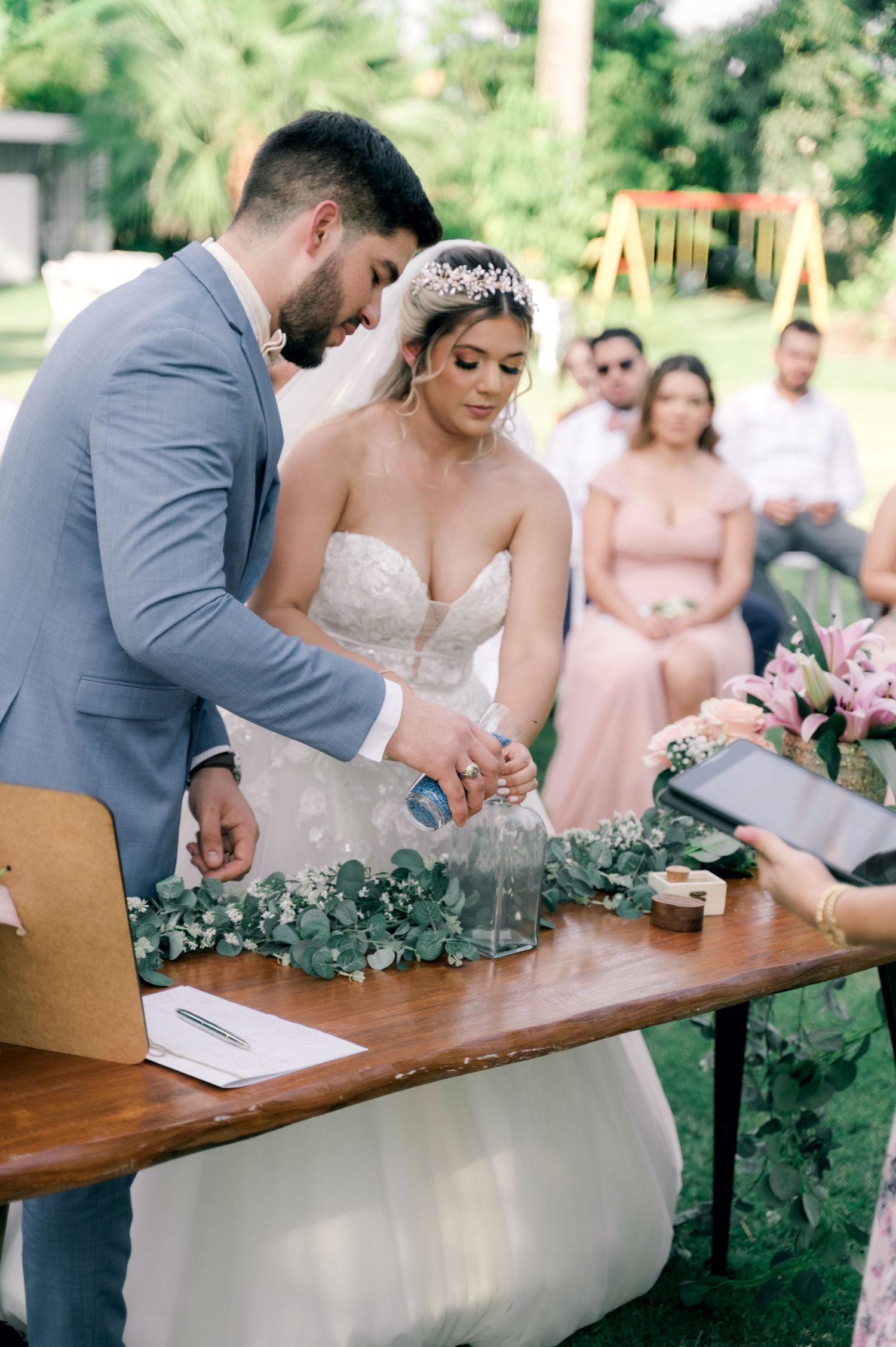 Wedding couple signs marriage license at wooden table outdoors surrounded by wedding party in pink and gray attire.