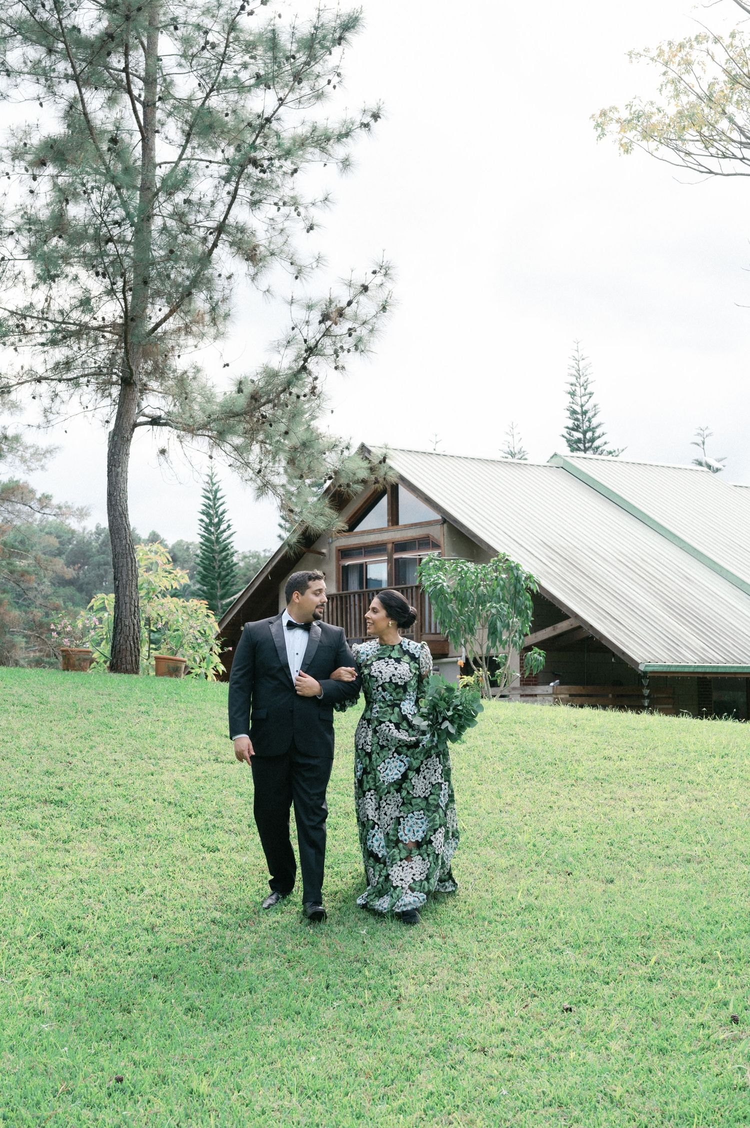 A couple walks together on a grassy hillside with pine trees and rustic buildings in the background.