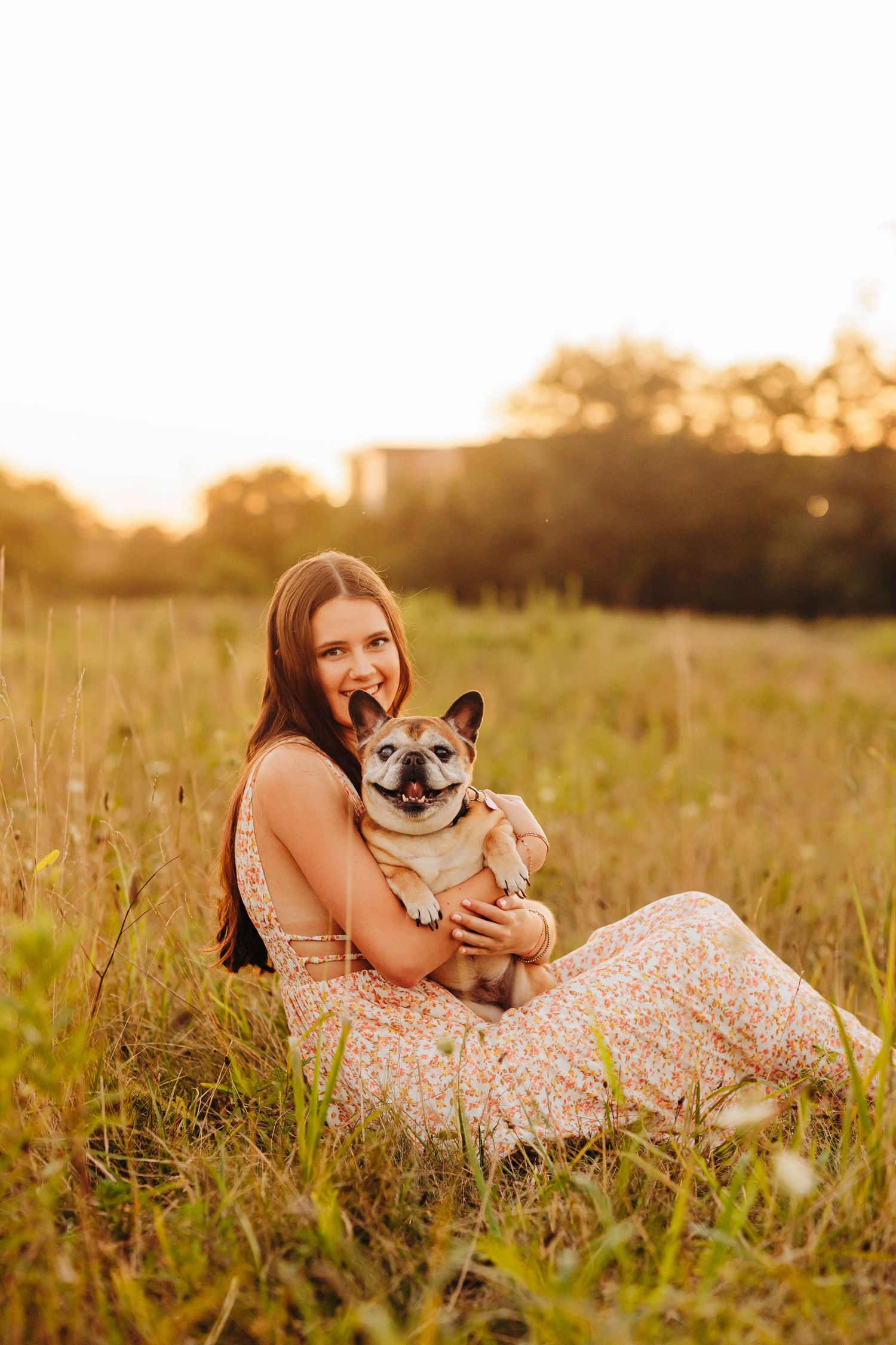 Woman in floral dress cuddles with smiling shiba inu dog in sunlit meadow.