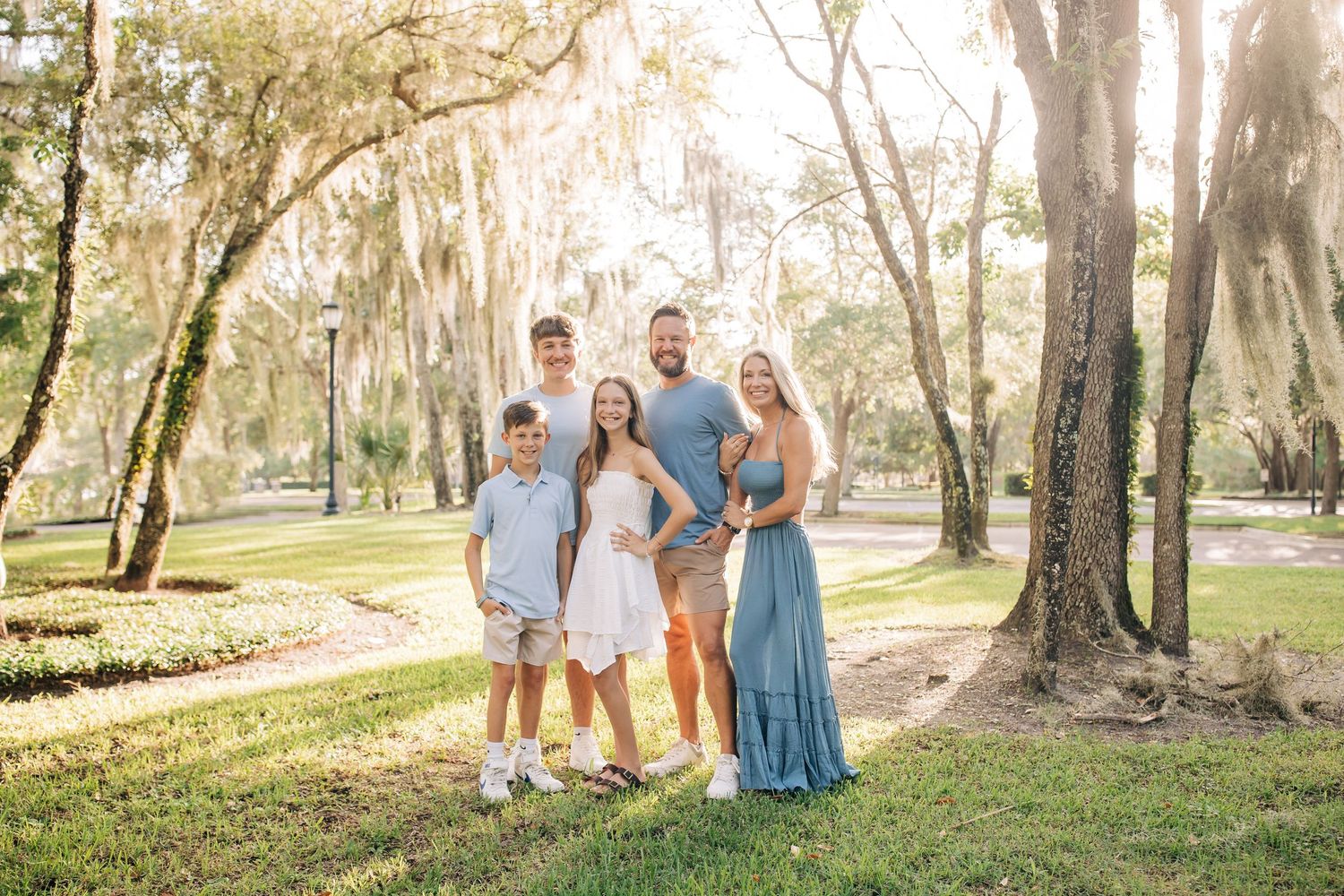 A joyful family of five poses together on a sunlit lawn surrounded by moss-draped oak trees.