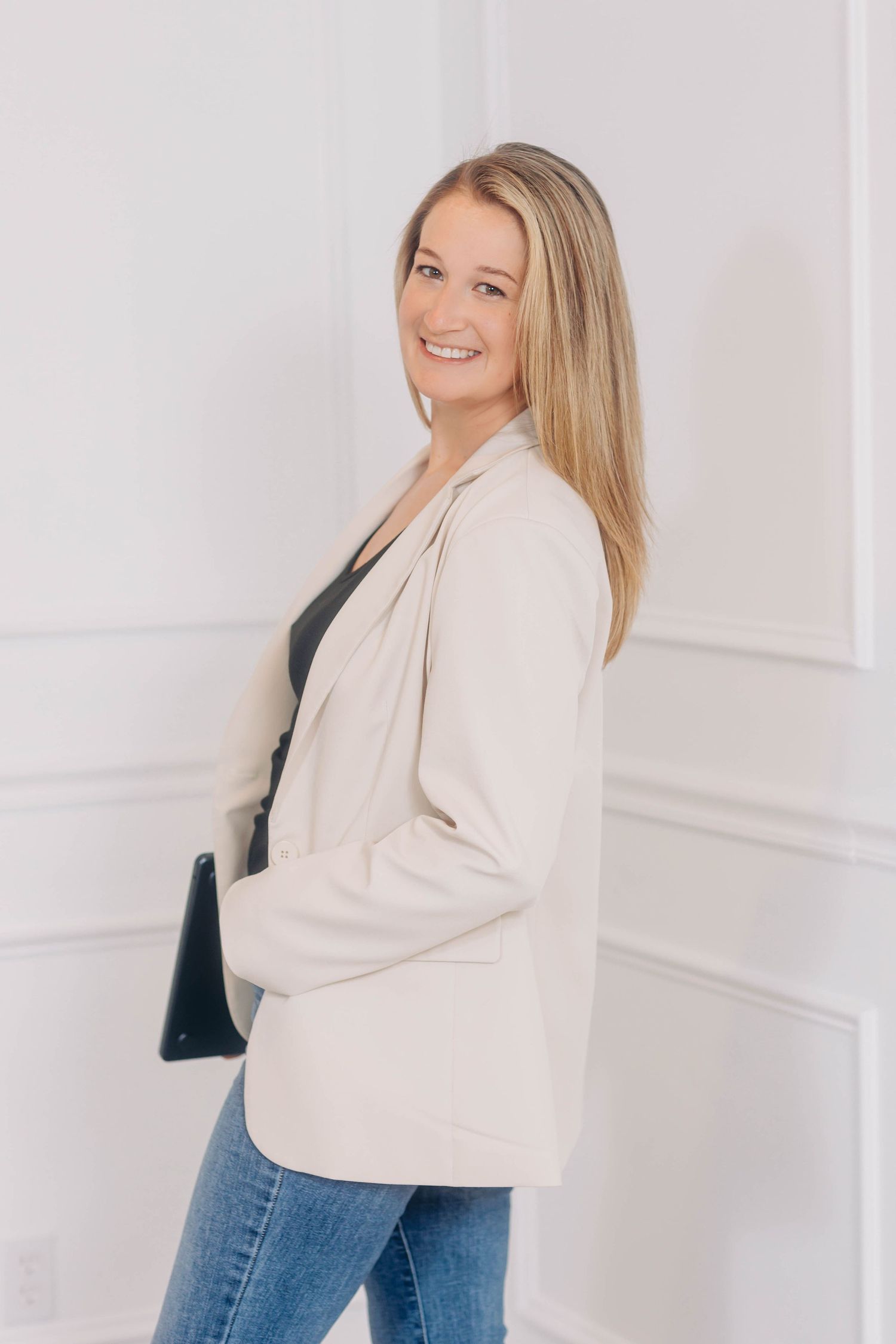 Woman in cream sweater and jeans poses against white wall in lifestyle photography session.