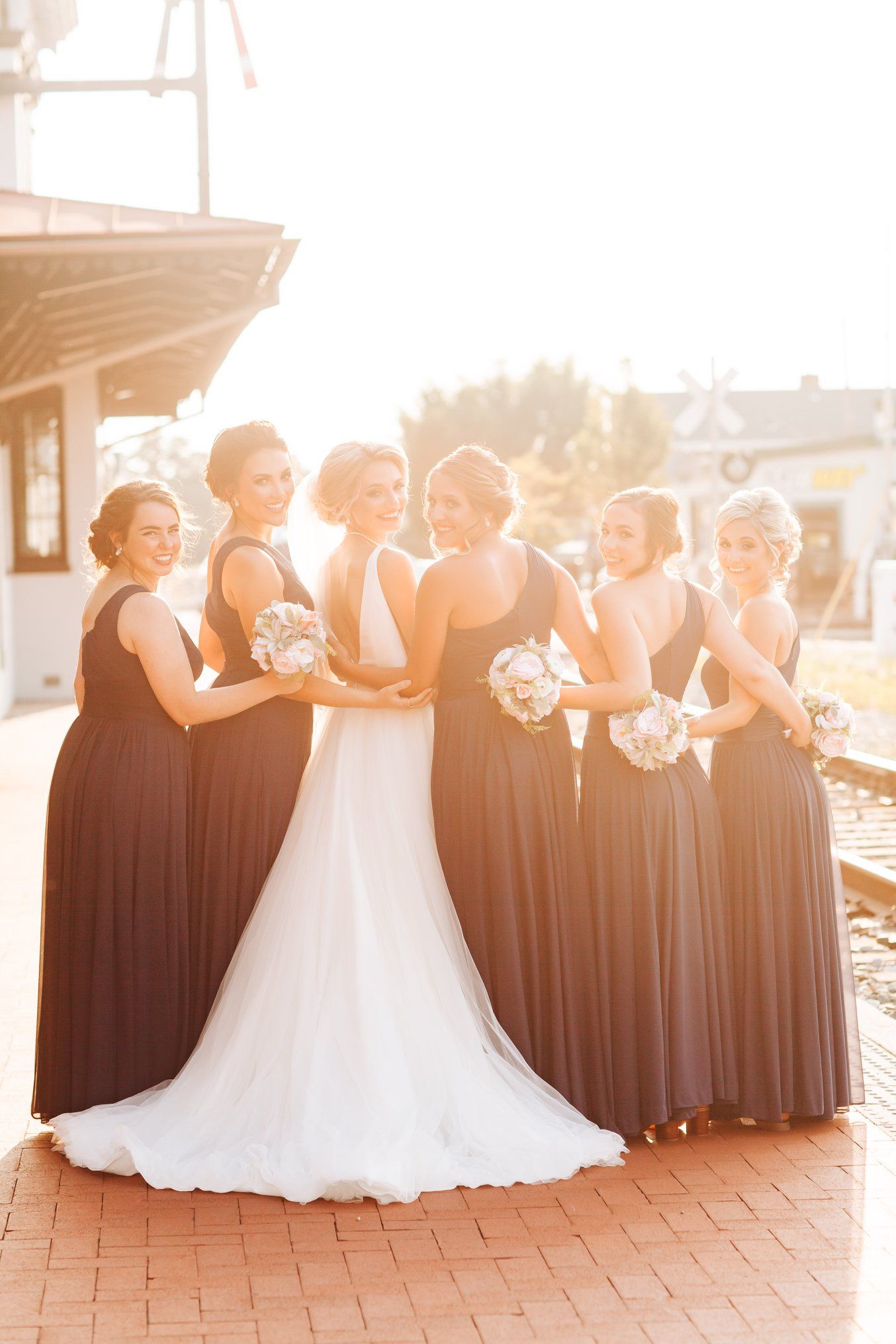 Wedding party in dark dresses poses together at sunset on brick patio.
